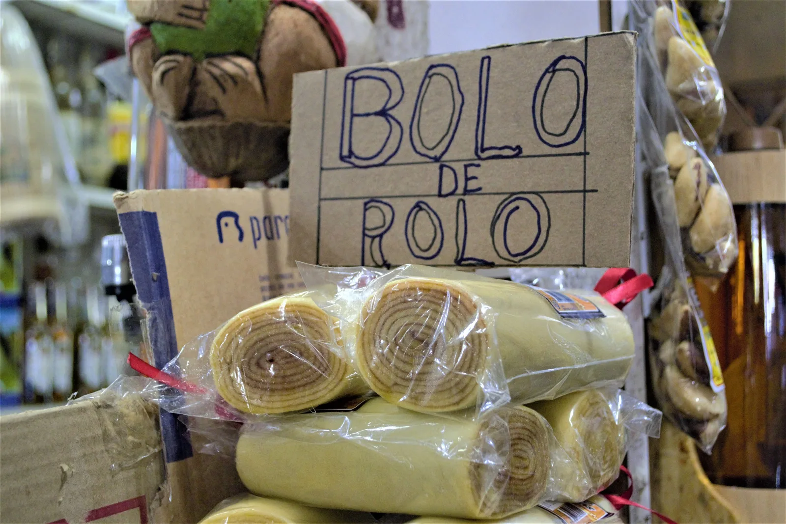 Rolled cakes wrapped in plastic stacked on a market stall under a handwritten cardboard sign reading Bolo de Rolo