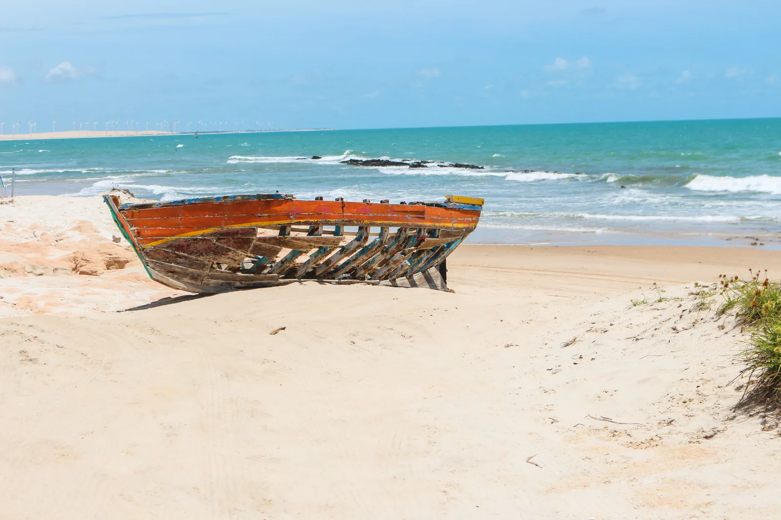The bare wooden ribs of a wrecked fishing boat lying half-buried on a pale beach at Canoa Quebrada, green sea and a low reef line behind
