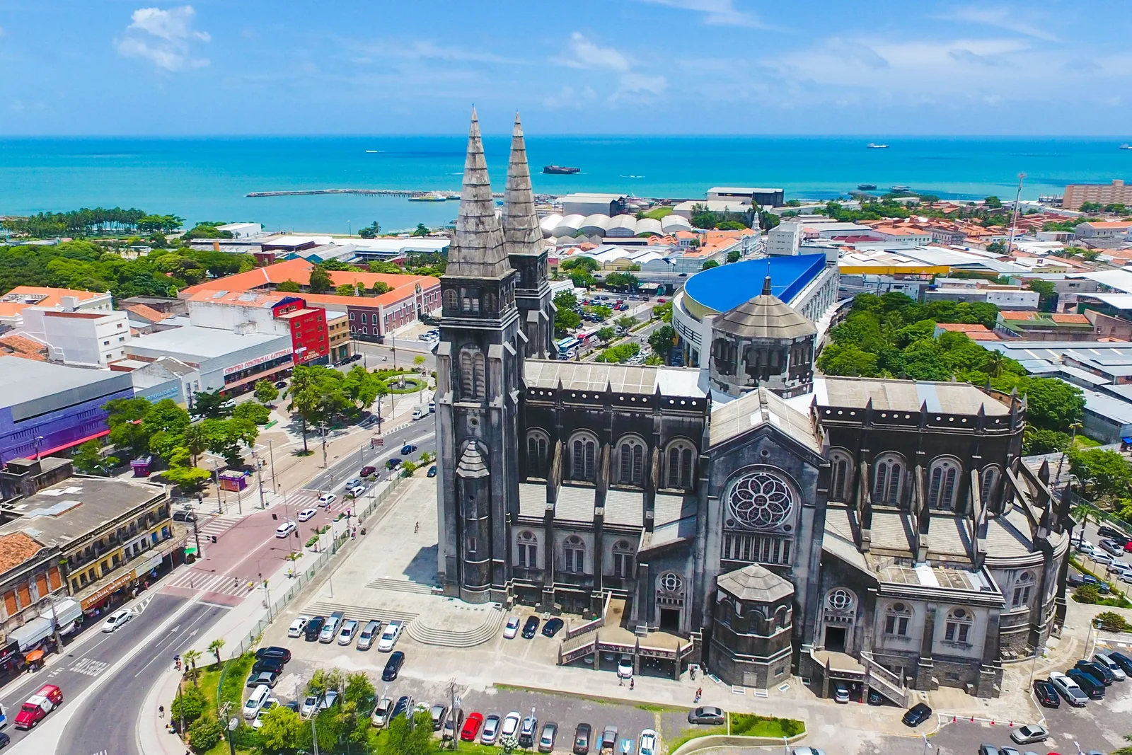 The twin gray spires of Fortaleza’s Catedral Metropolitana from above, with the blue curved roof of the Mercado Central beside it and the sea on the horizon