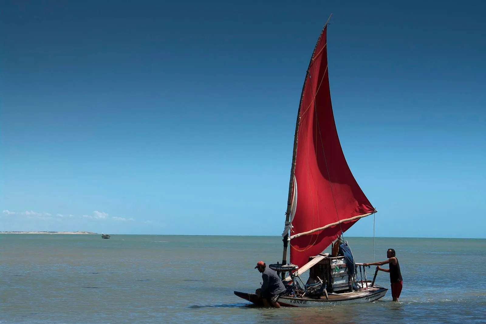 A jangada under a deep red sail on flat shallow water at Flecheiras, two men aboard and one standing waist-deep beside it