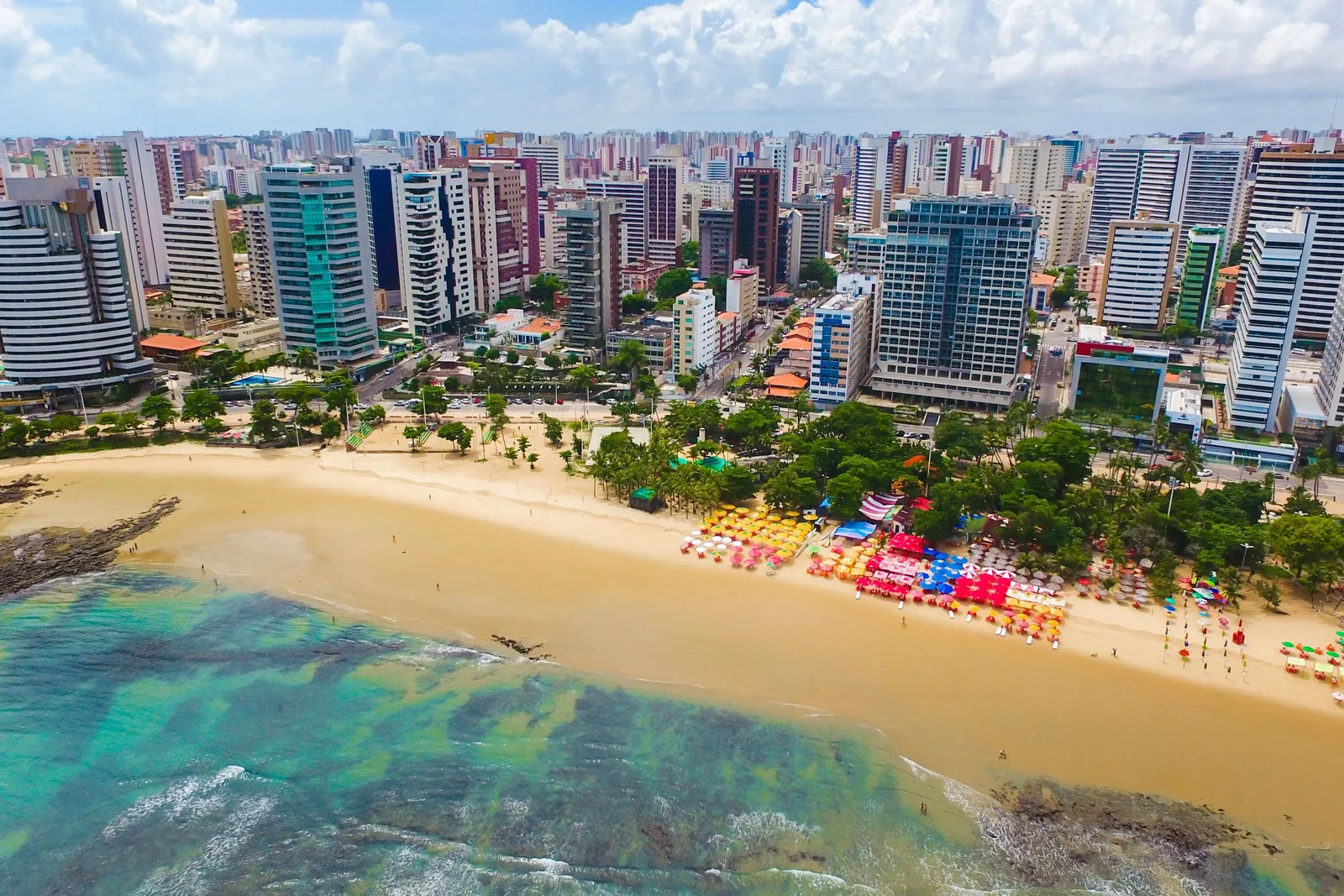 Meireles beach from the air: turquoise shallows over pale sandbanks, a beach packed with rows of colored parasols, and the unbroken wall of apartment towers behind it