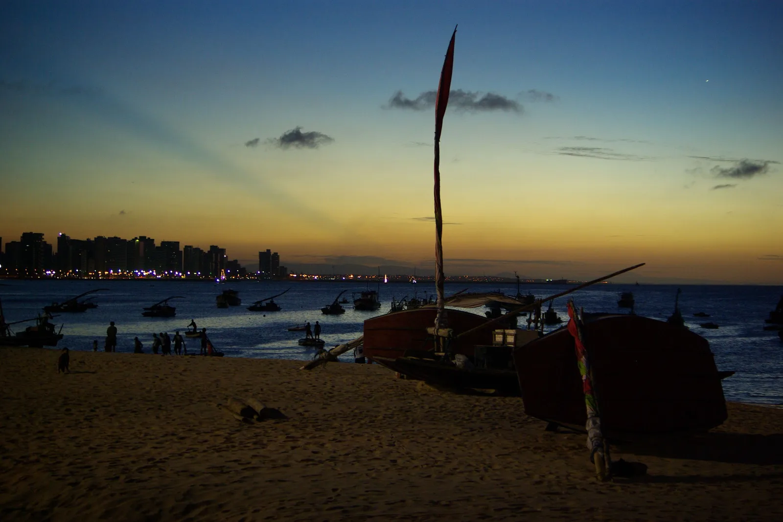Dusk at Mucuripe: a jangada silhouetted on the sand with its mast against a gold and blue sky, and the lights of the city strung along the shore behind