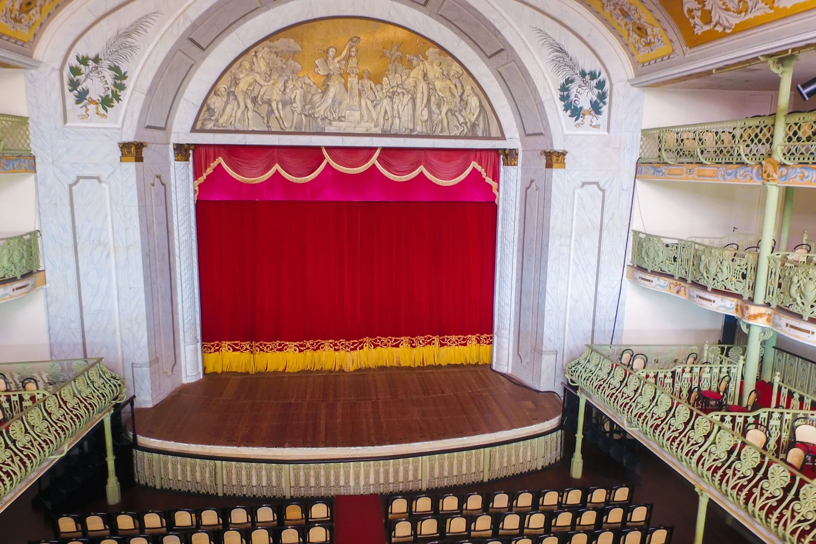 The stage of the Theatro José de Alencar seen from a side box: a red curtain under a painted arch, with tiers of cast-iron balconies and rows of wooden seats below