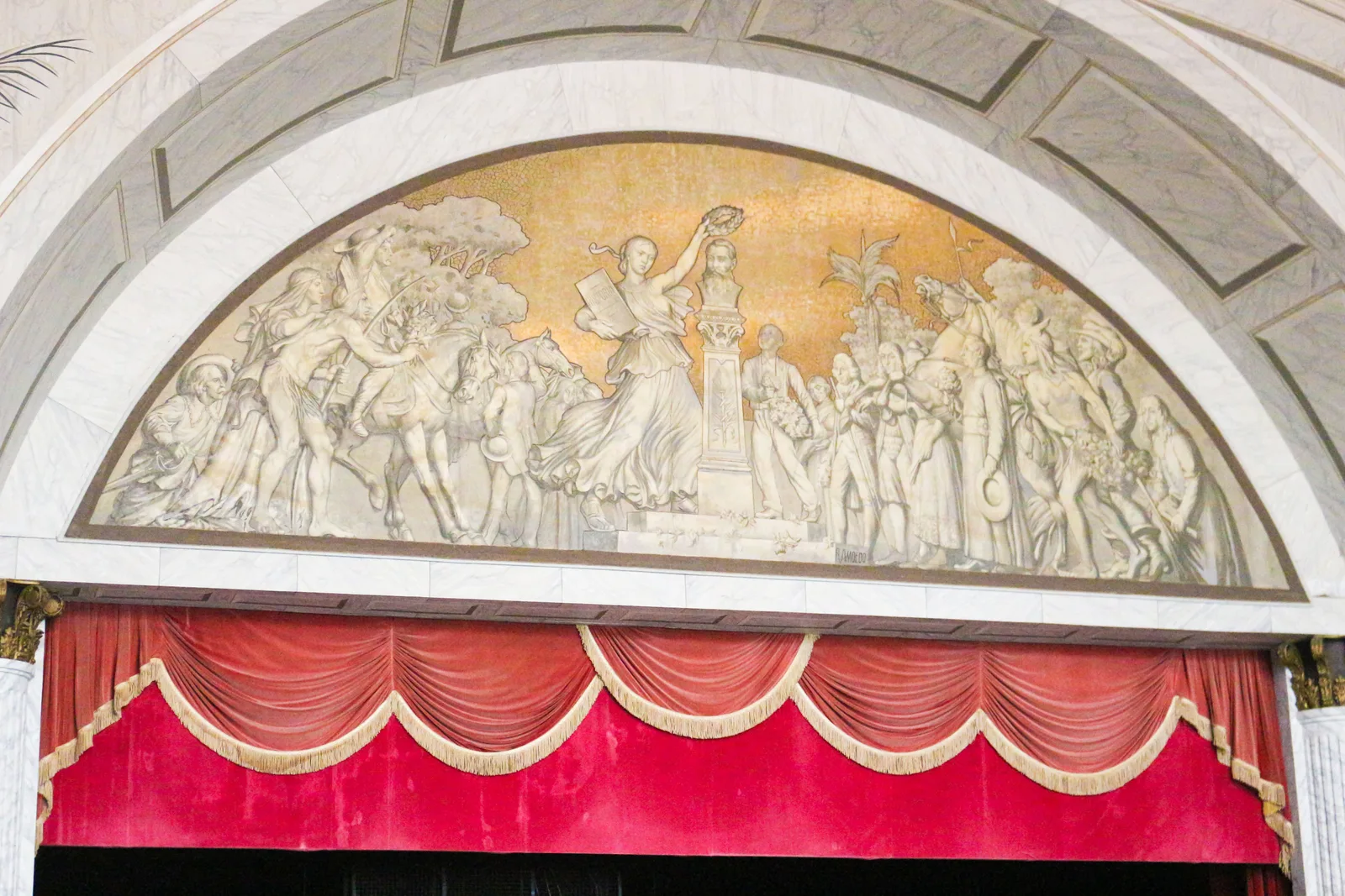 A pale plaster relief of classical figures set into the arch above the Theatro José de Alencar’s stage, with the red and gold curtain drawn beneath it