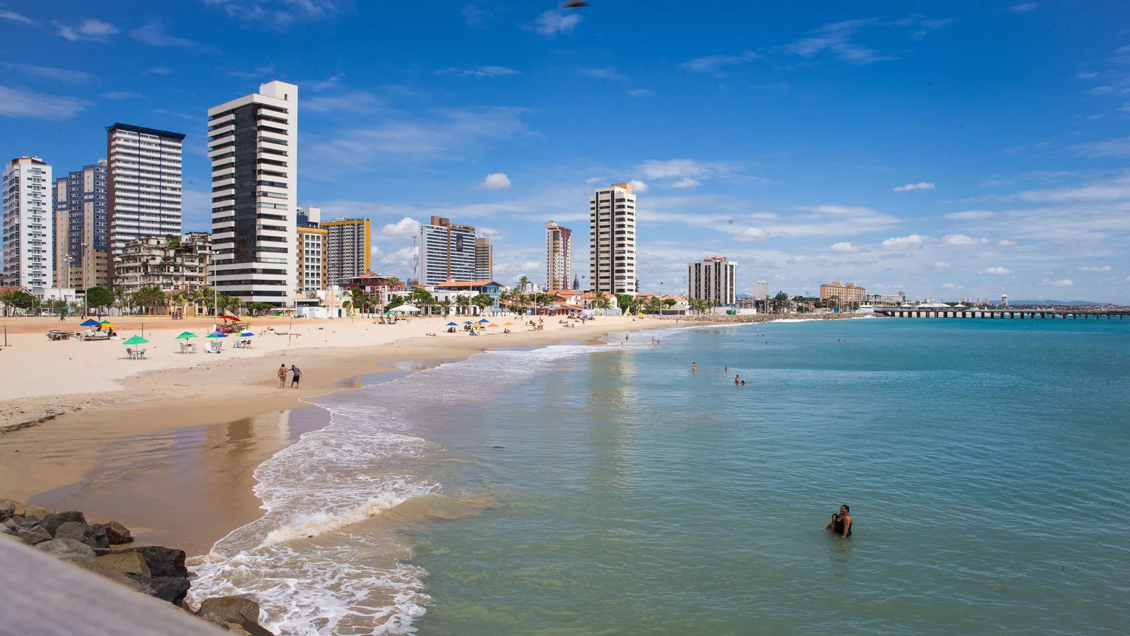 The seafront at Praia de Iracema in Fortaleza: a curve of pale sand under a line of apartment towers, colored beach parasols along the back of the beach and a long pier running out into green-blue water