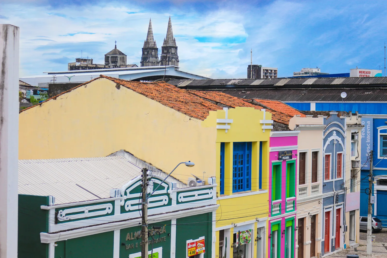A row of colorful colonial shopfronts in Fortaleza’s old center painted yellow, green, pink and blue, with the twin gray spires of the cathedral rising behind the roofs