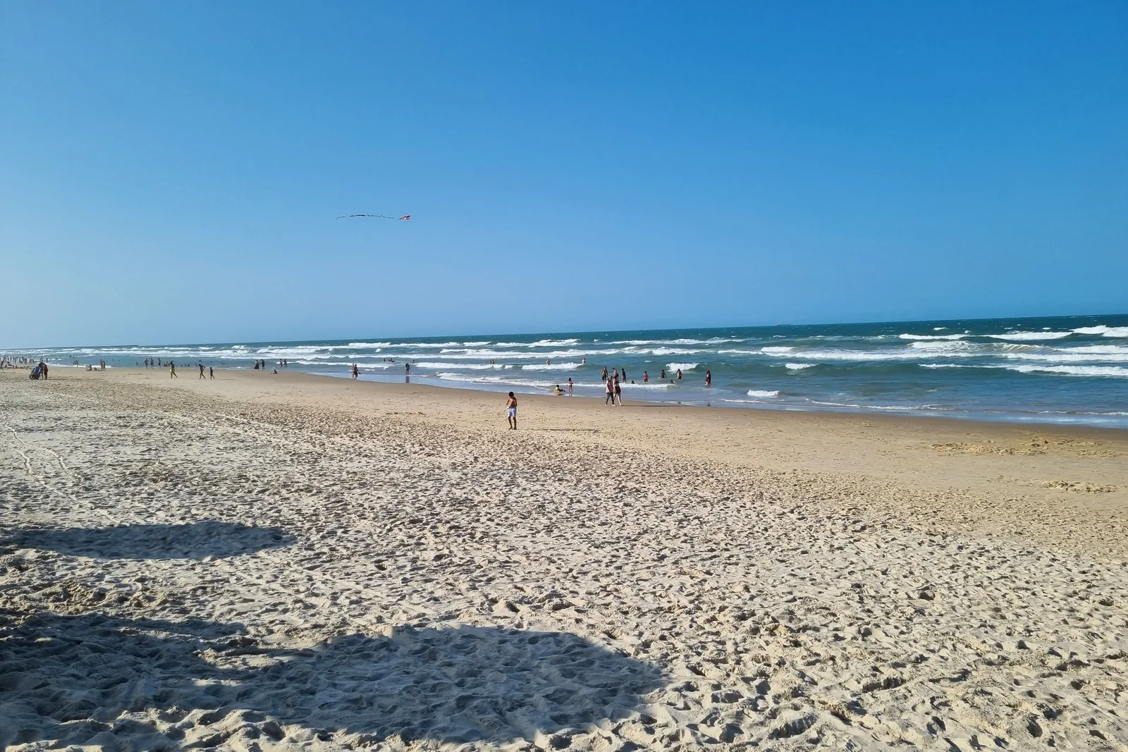 A wide sandy beach at Praia do Futuro with white surf running up it, a scattering of people at the waterline and a bright hazy sky over the sea