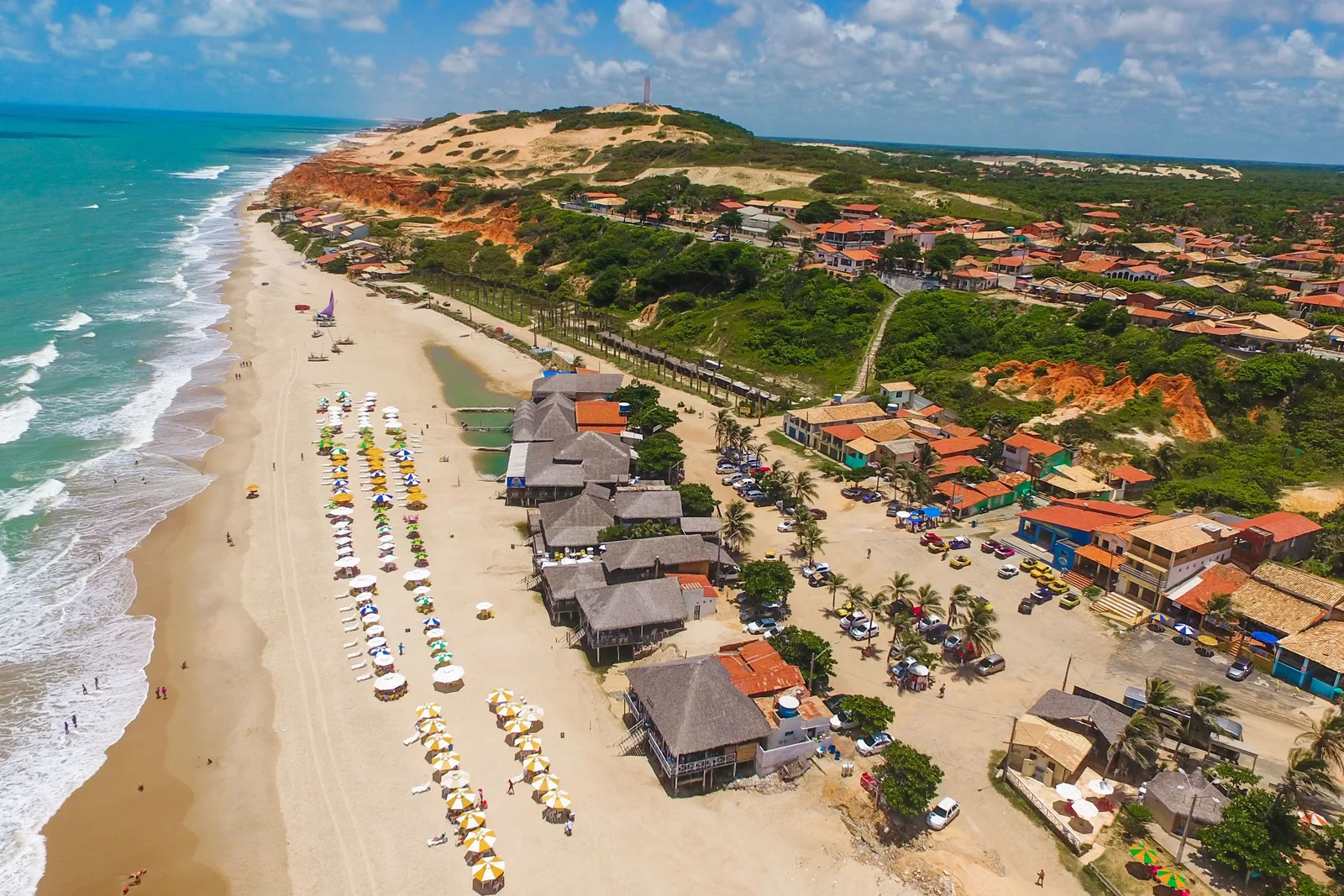 Morro Branco from the air: a wide beach with thatched barracas and a line of jangadas on the sand, red and cream cliffs rising behind them and pale dunes beyond