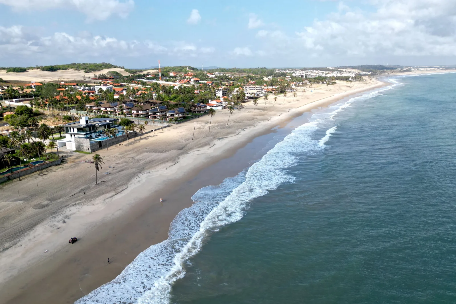 The coast west of Fortaleza from the air: a long curve of pale beach with surf breaking along it, and a village of low buildings, red-roofed villas and palms set back behind the sand with dunes rising beyond