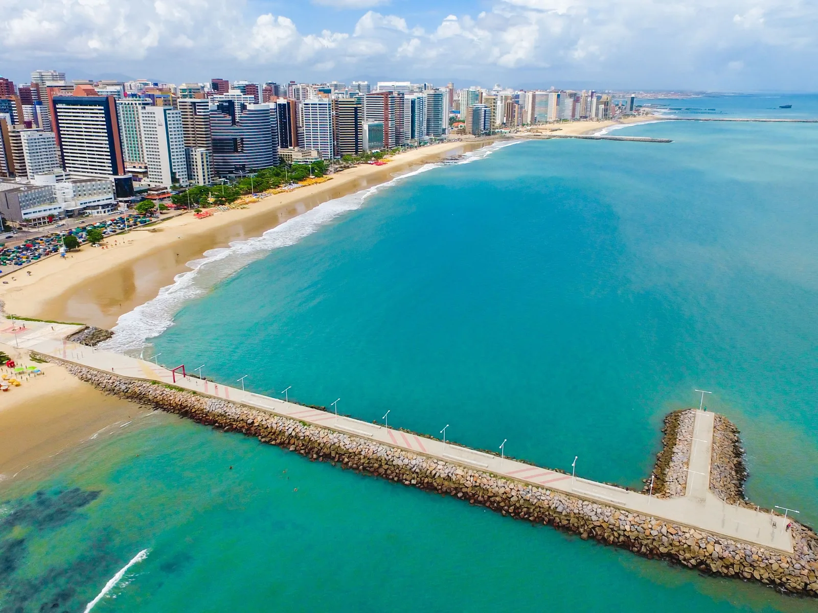 The Meireles seafront from the air: a wall of apartment towers along a curve of pale sand, and a long stone breakwater with a lit walkway on top running out into flat turquoise water