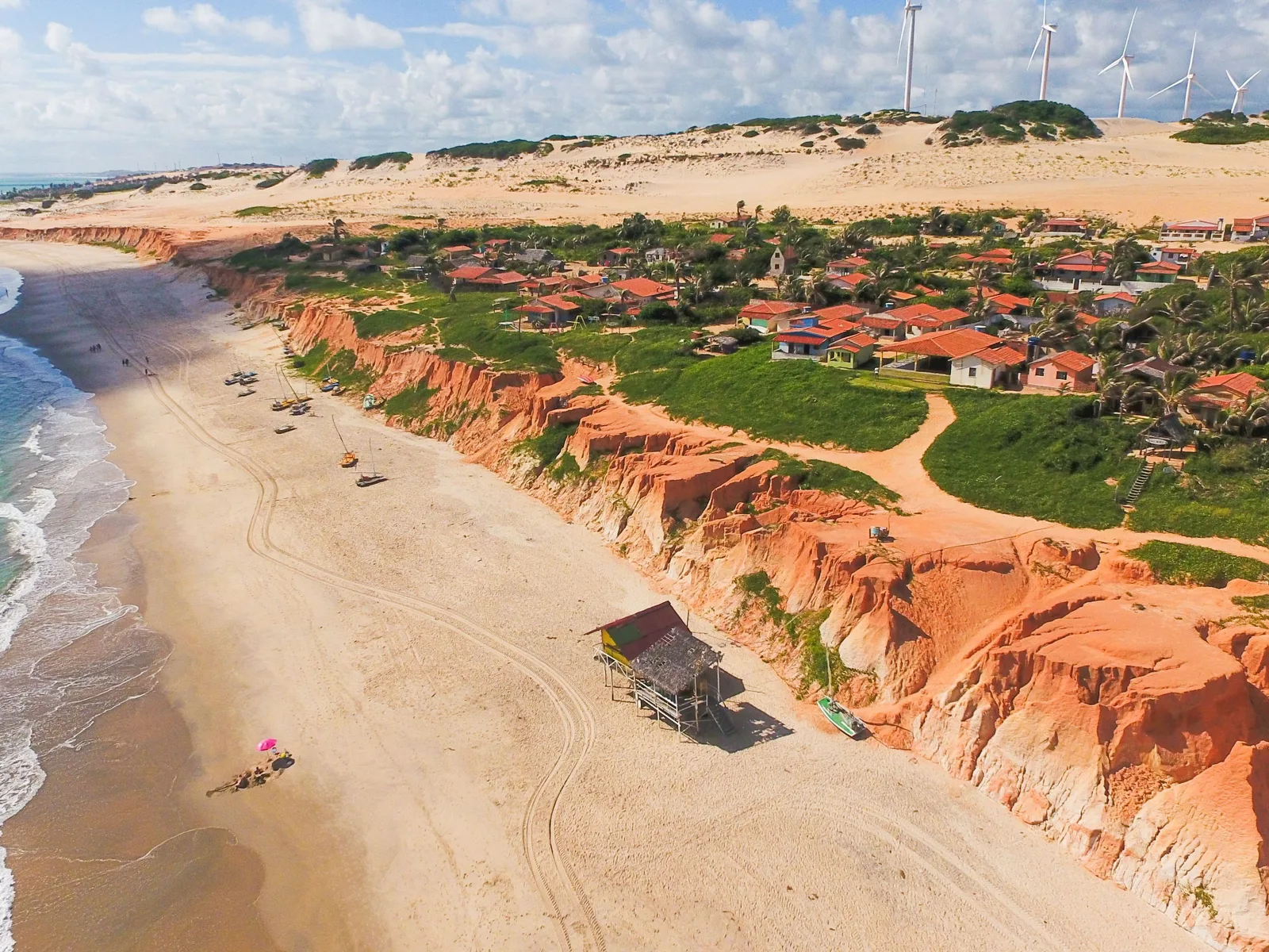 Canoa Quebrada from the air: red and cream sandstone cliffs dropping to a wide pale beach with jangadas and buggy tracks on it, red-roofed houses on the green clifftop, and pale dunes with a line of wind turbines behind them
