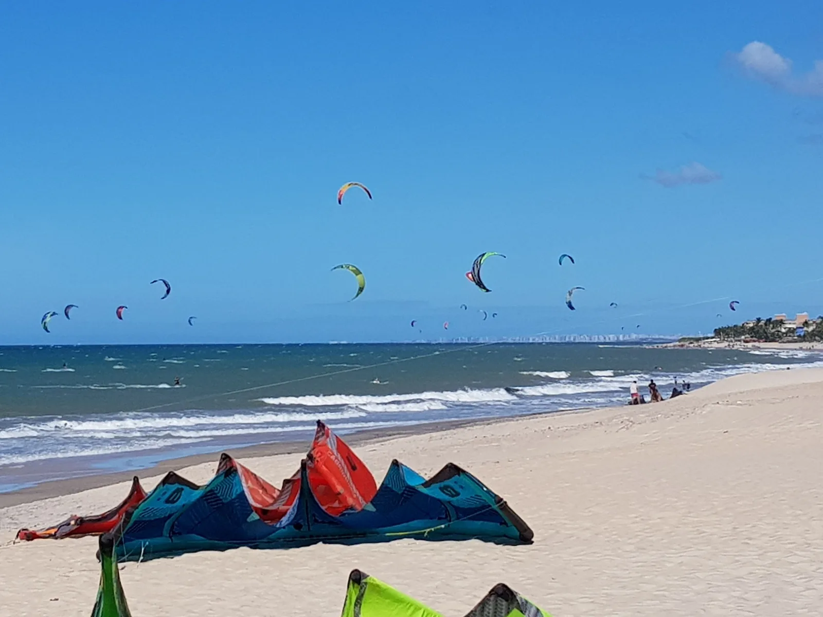 The beach at Cumbuco with a dozen kitesurfing kites in the air over the sea, more kites laid out on the sand in the foreground, and the faint outline of Fortaleza’s towers on the horizon