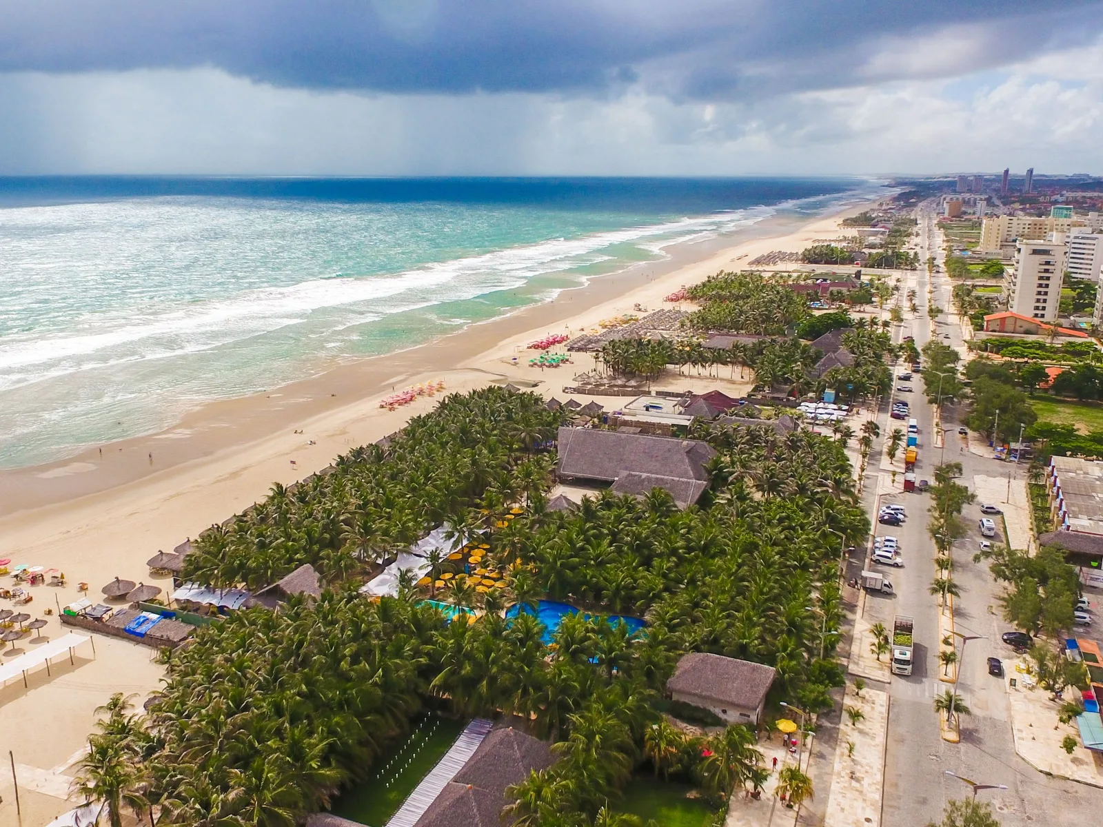 Praia do Futuro from the air: surf breaking along a wide pale beach, a run of thatched barraca roofs and coconut groves set back from the sand with pools and yellow parasols among them, the avenue behind, and a rain squall out over the sea