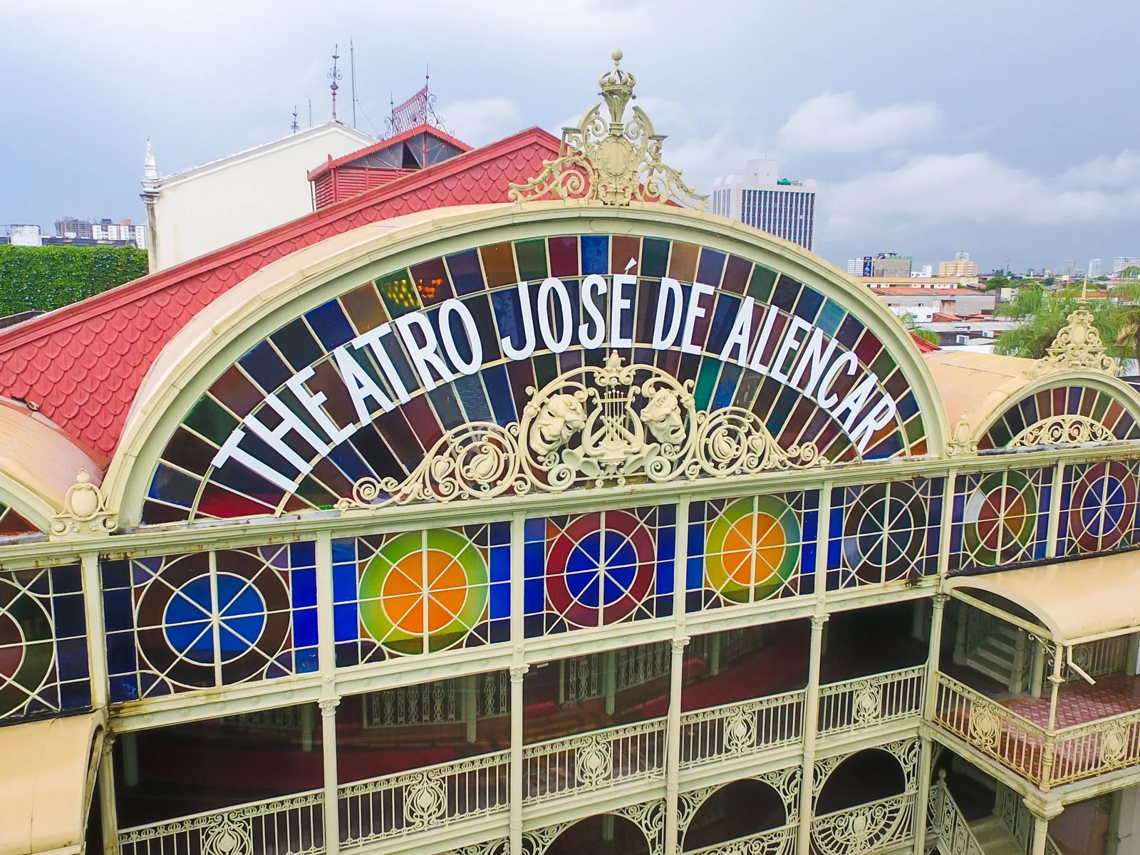 The front of the Theatro José de Alencar in Fortaleza: a pale green cast-iron arch filled with panels of colored stained glass, the theater’s name arched across it in white capitals, with a red tiled roof behind and city towers beyond