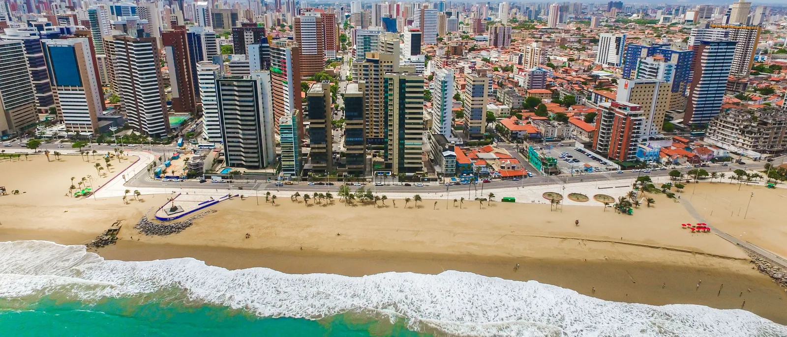 Praia de Iracema from above in a wide frame: a beach and rock groynes in the foreground with a dense line of apartment blocks and hotel towers running along behind it