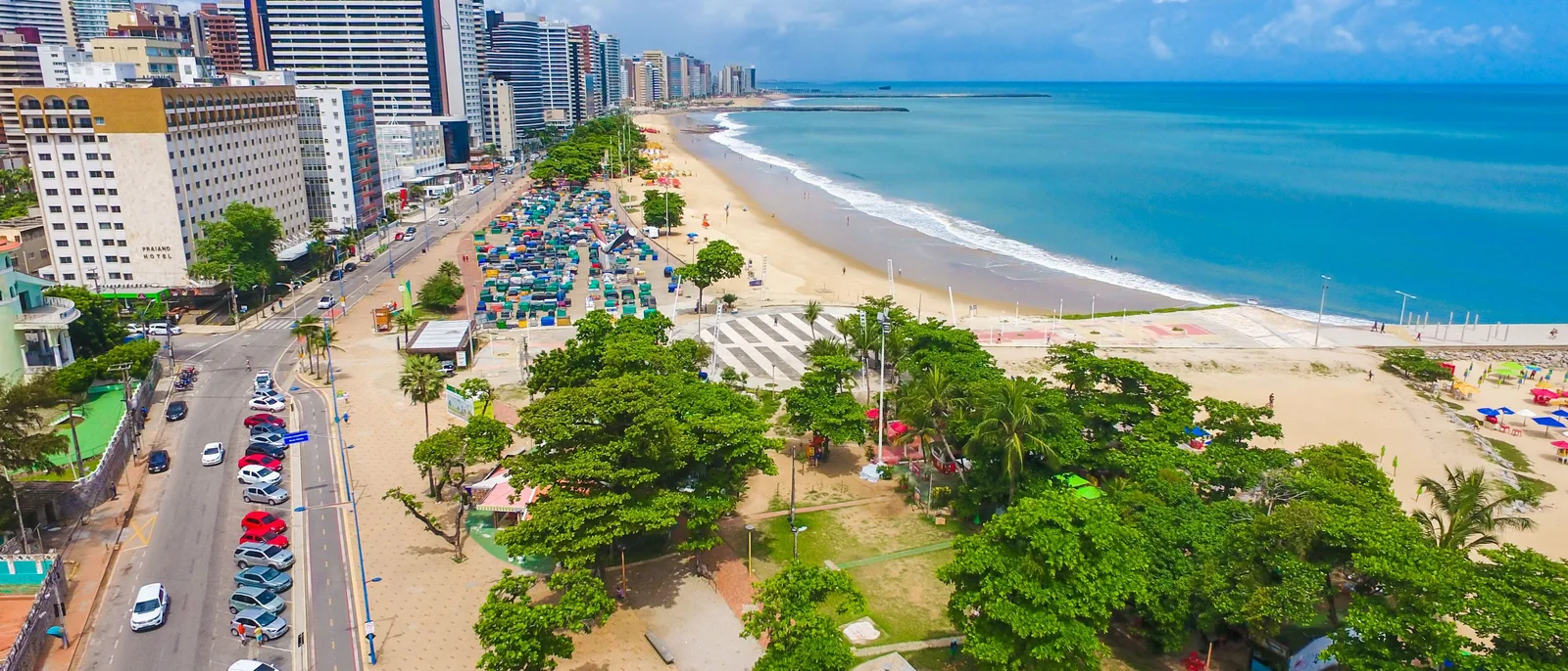 The Meireles seafront in a wide frame: a beach lined with colored parasols, a green promenade with palms along it, and a solid line of apartment towers behind, with turquoise water in front