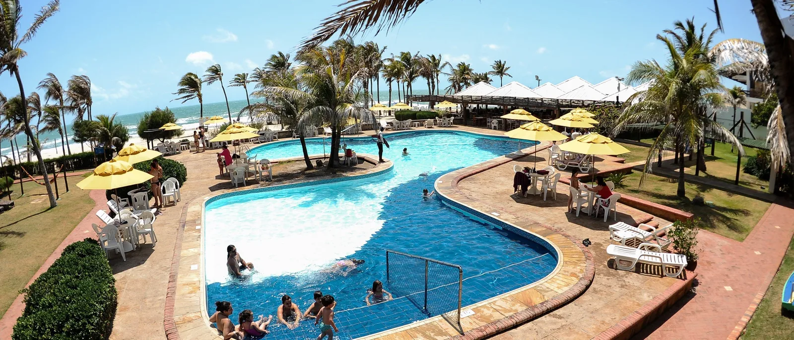 A resort swimming pool at Porto das Dunas in a wide frame: a curving pool with people in it, yellow parasols and loungers along the tiled edge, and coconut palms behind