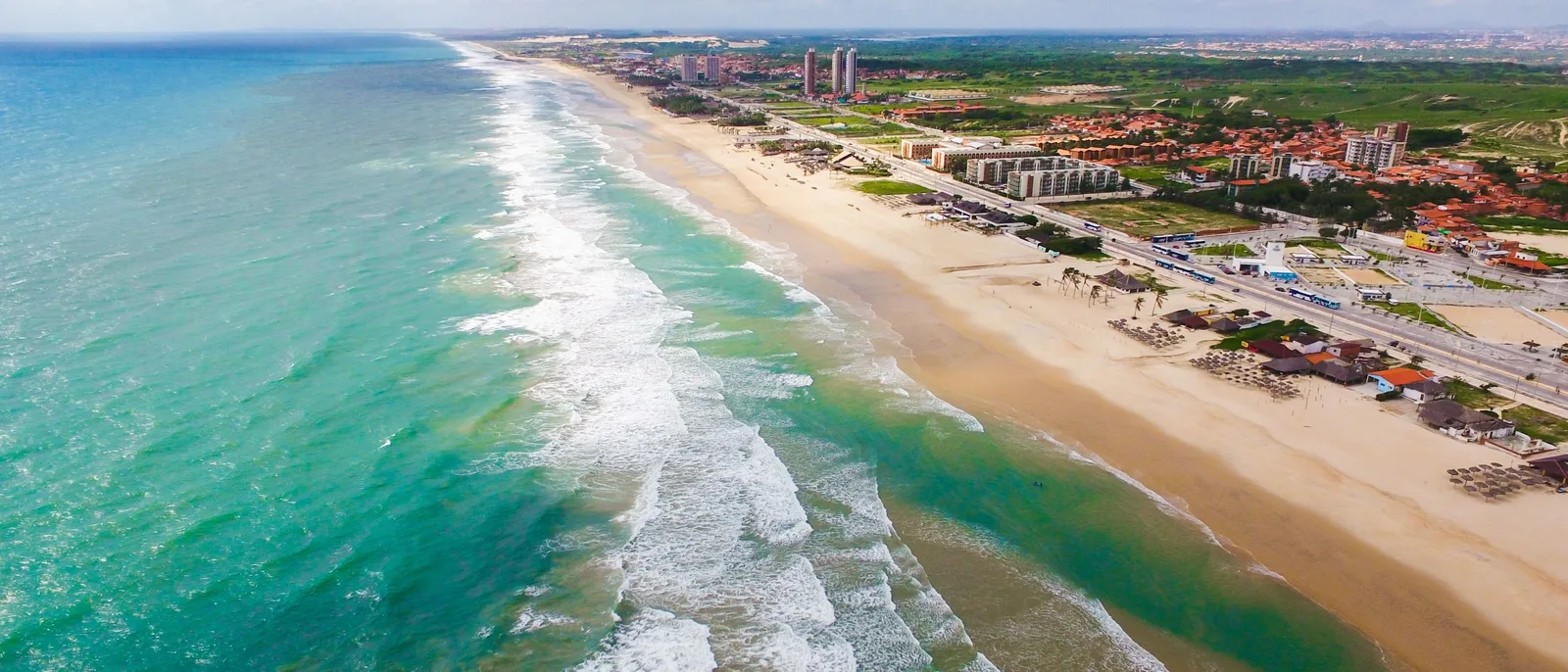 Praia do Futuro from the air in a wide frame: surf running along a broad pale beach with thatched barracas and coconut groves behind it and the city towers far off along the shore