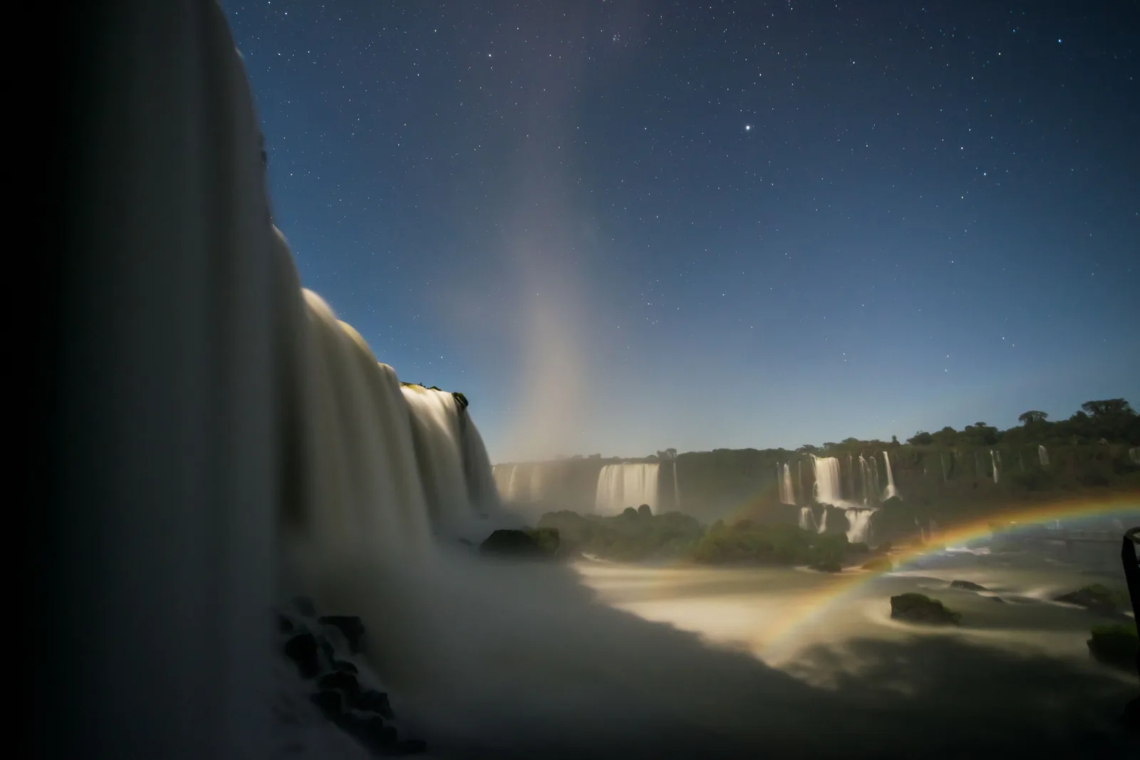 Iguazu Falls at night under a full moon, stars over the gorge and a moonbow arcing through the spray at the foot of the falls