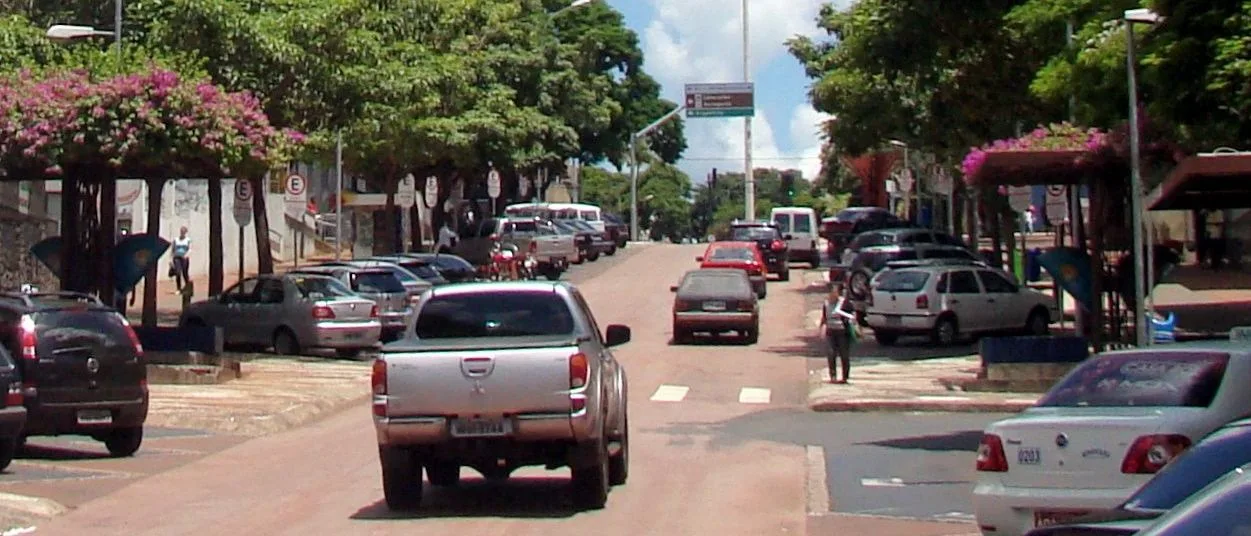 Avenida Brasil in central Foz do Iguaçu, cars parked along both sides, shopfronts under the trees and traffic running away toward the end of the street