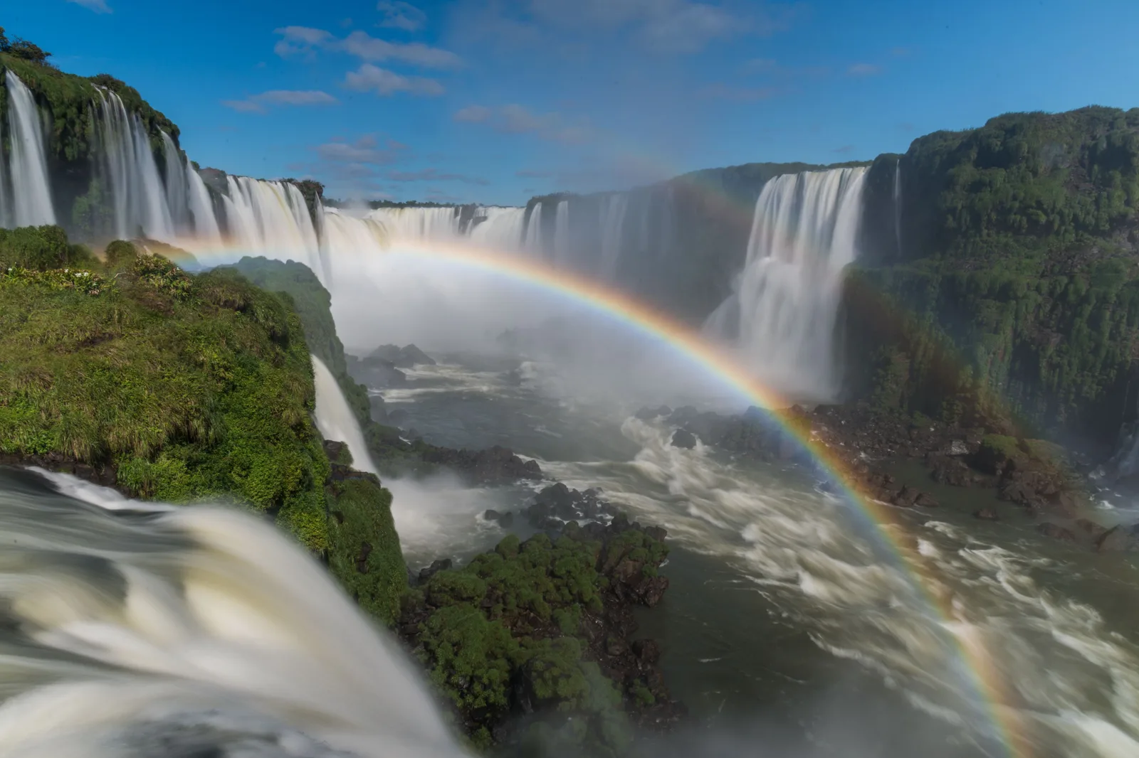 A rainbow arching over the Iguaçu falls, water pouring down between green ledges