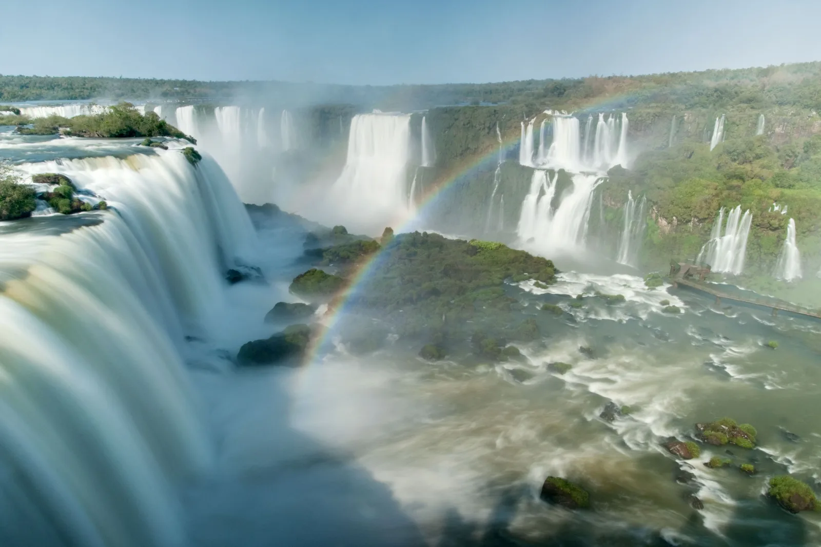 A wide view of a row of waterfalls with a rainbow in the spray at their feet