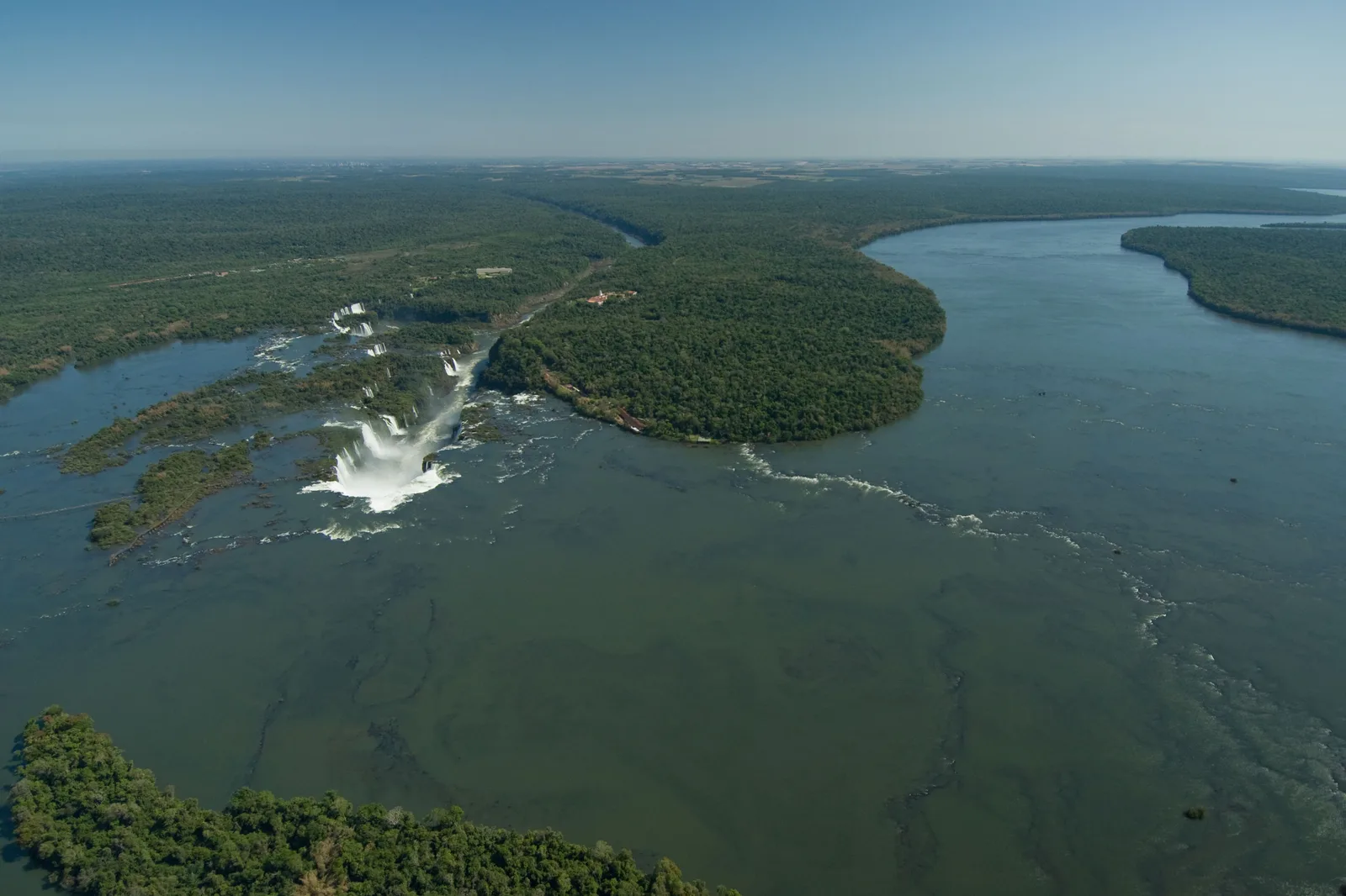 The wide Iguaçu river from high above, the falls a white line across it and rainforest to the horizon