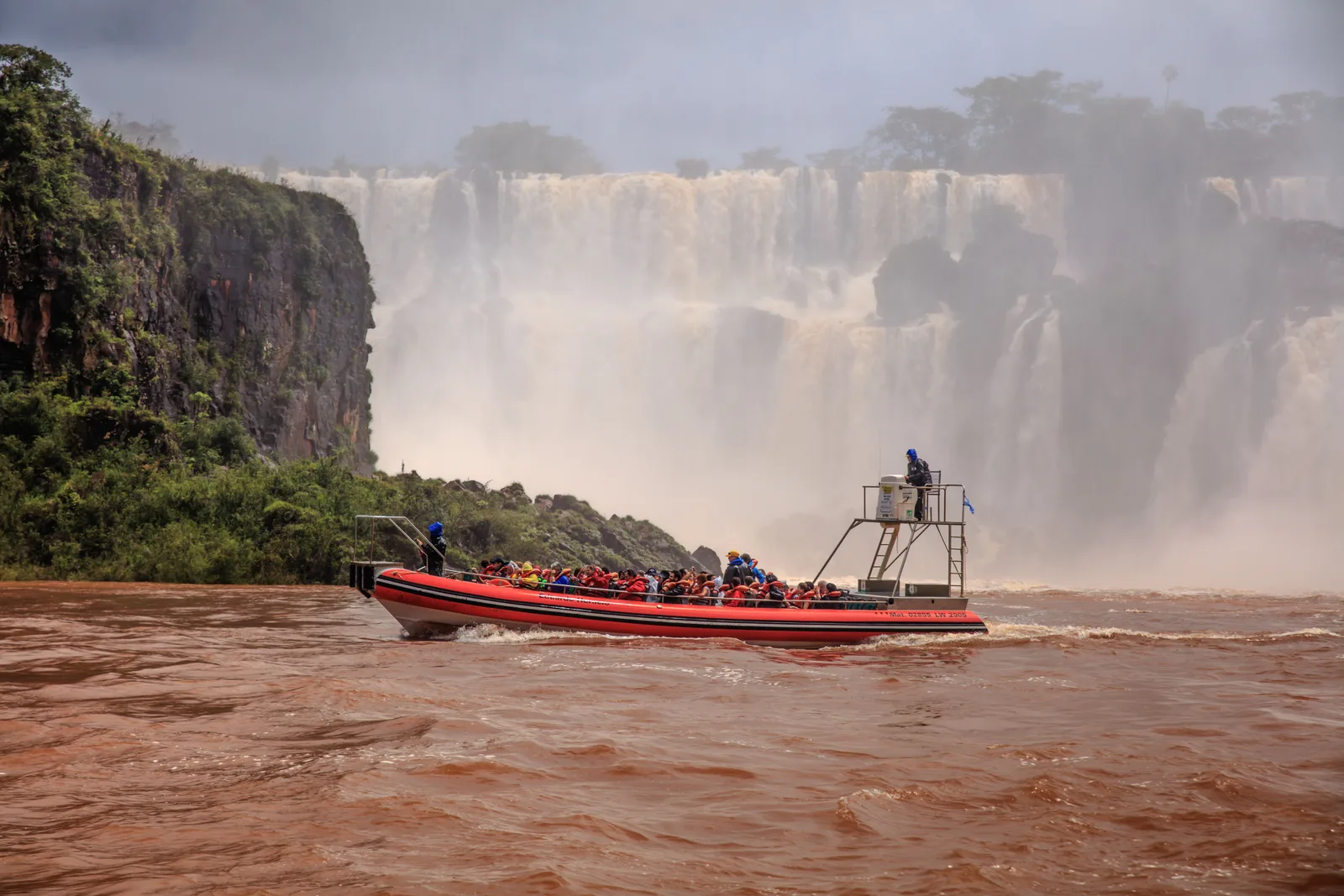 A boatload of passengers on the brown river below the falls on the Argentine side, a wide wall of water coming down behind them