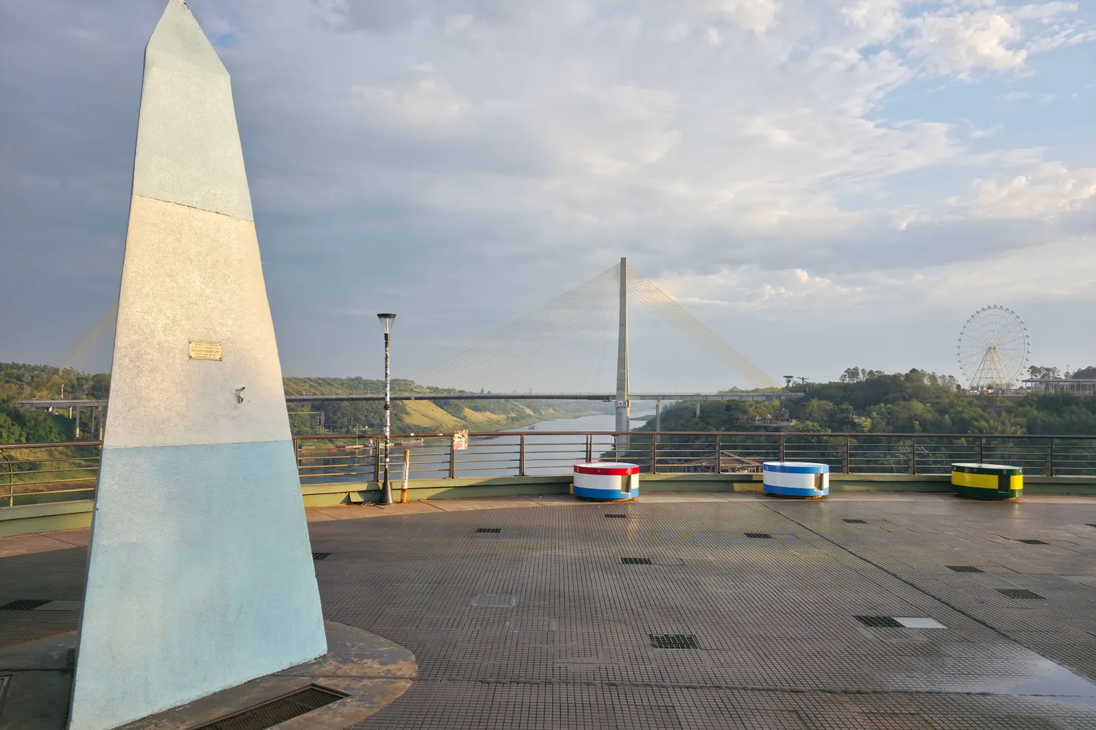The pale blue and white Argentine obelisk at the Hito Tres Fronteras, standing on an empty paved terrace above the river with a bridge in the distance behind it