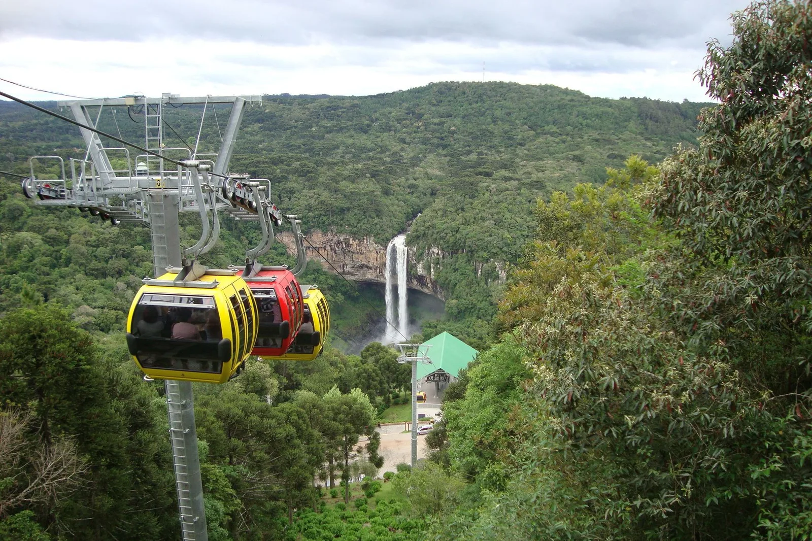 Yellow and red cable-car cabins hanging from a cable over the forest, with the Caracol waterfall falling from a cliff in the distance behind them