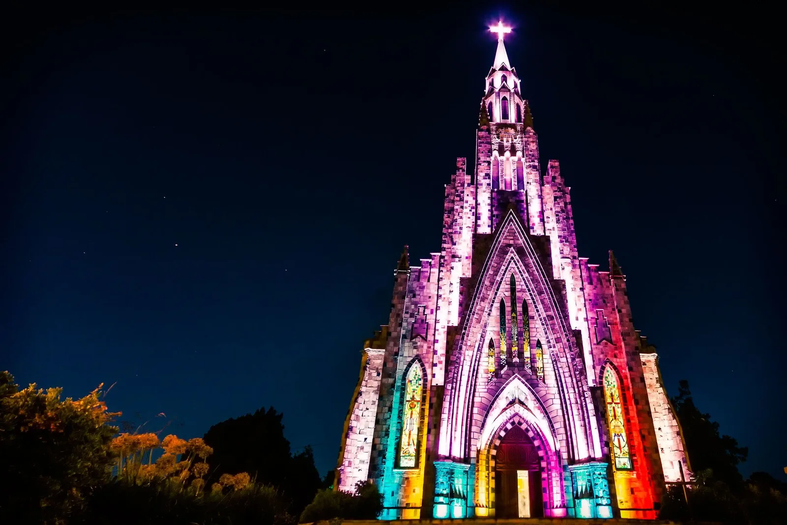 The Catedral de Pedra at night, its Gothic façade and spire lit in pink, violet and gold against a dark sky