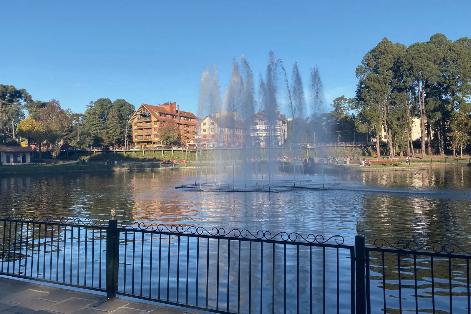 The Lago Joaquina Rita Bier on a sunny day: fountains jetting up from the middle of the lake, a black railing in the foreground and hotels among tall trees on the far side