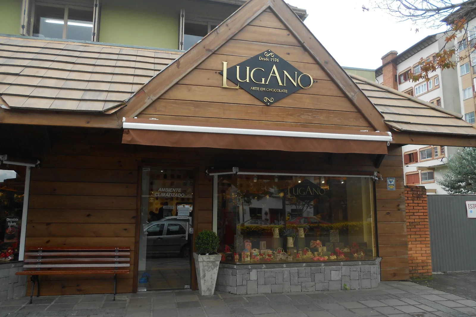 The front of a Lugano chocolate shop in Gramado: a wooden chalet-style gable with the Lugano name over a window full of boxed chocolates, a bench beside the door