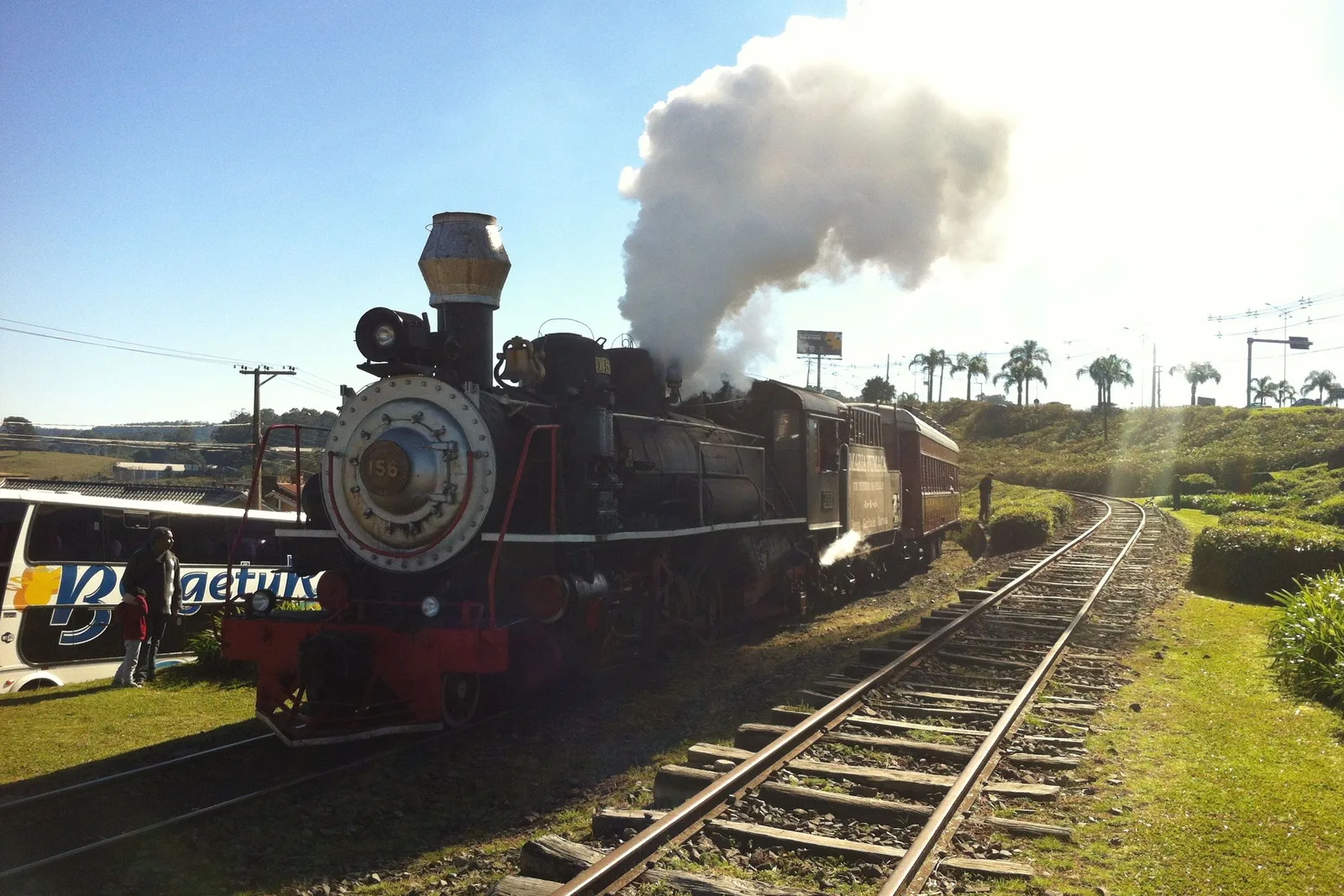 The Maria Fumaça steam locomotive standing on the track at Carlos Barbosa with white smoke pouring from its chimney into a bright sky