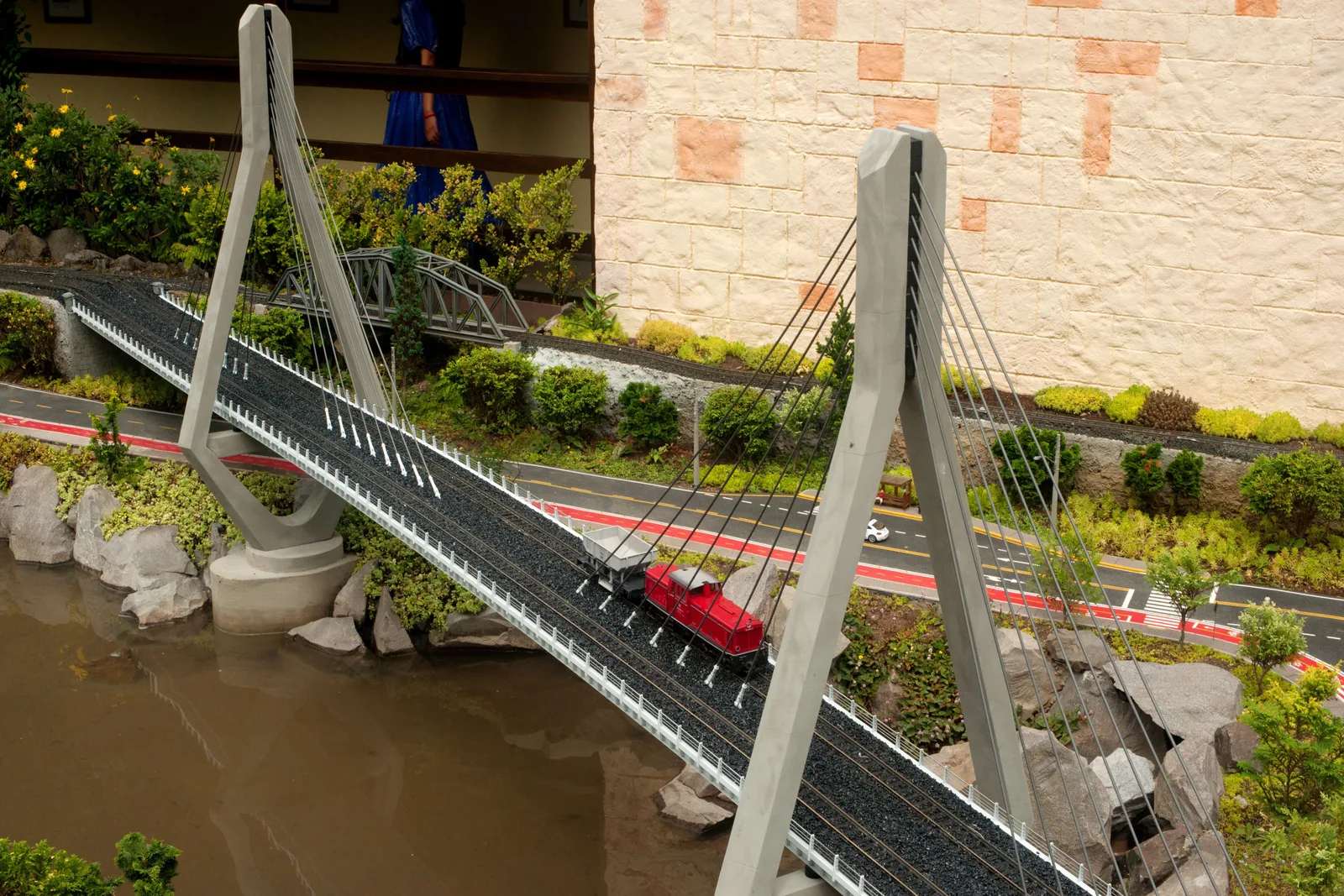 A model cable-stayed bridge at Mini Mundo with a small red train crossing it over a pond, model roads and planted rockwork around it