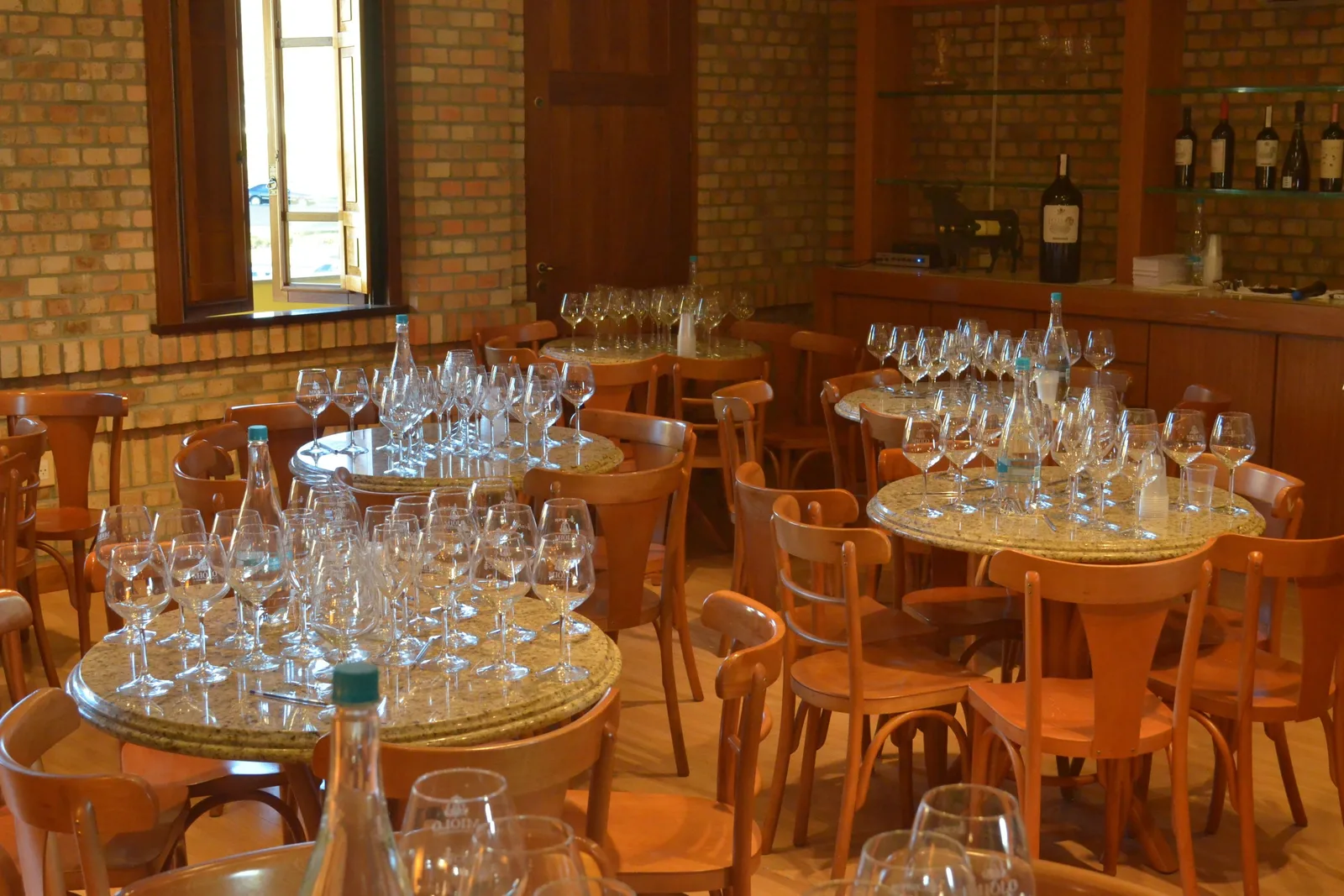 A tasting room at Miolo laid for a group: round tables covered with rows of empty wine glasses, wooden chairs and a brick wall with a window behind