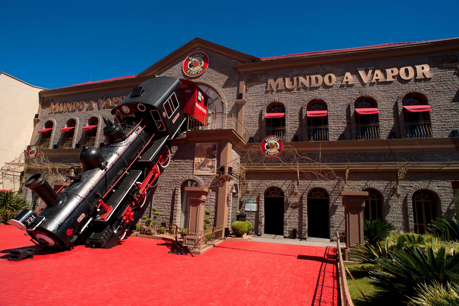 The Mundo a Vapor building in Canela: a stone façade with red awnings and a full-size black steam locomotive set at an angle as if crashing through the front, on a red forecourt