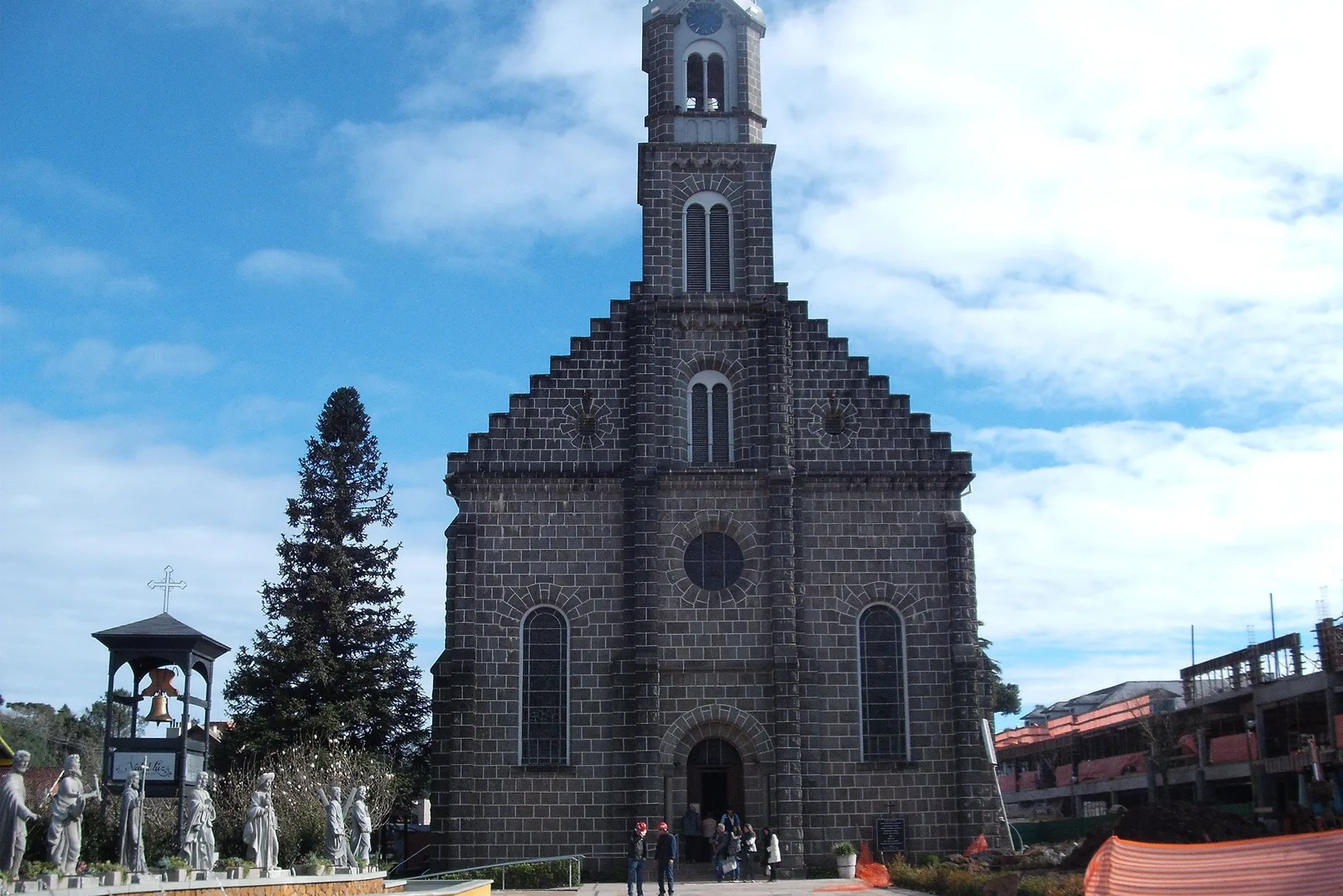The Igreja São Pedro in Gramado: a dark stone church with a tall square tower under a blue sky with cloud, a pine tree and white statues on the left