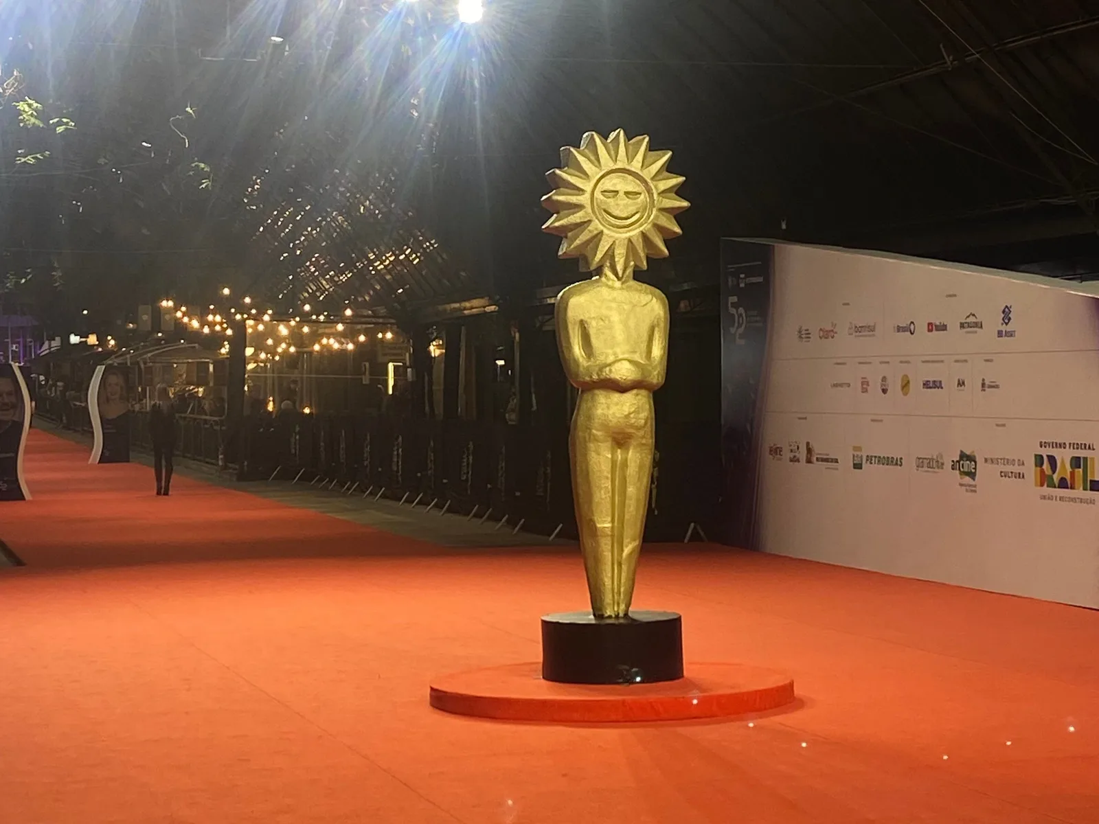 A tall golden Kikito, the sun-headed statuette created by the artisan Elisabeth Rosenfeld, standing on the red carpet at the entrance to the Gramado film festival, lights strung overhead