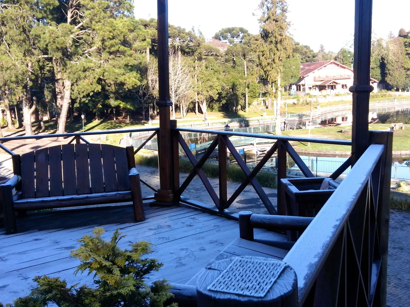 White frost on the boards, a bench and the wooden railings of a lakeside deck at the Lago Joaquina Rita Bier on a sunny morning, with tall pines and a red-roofed building across the water