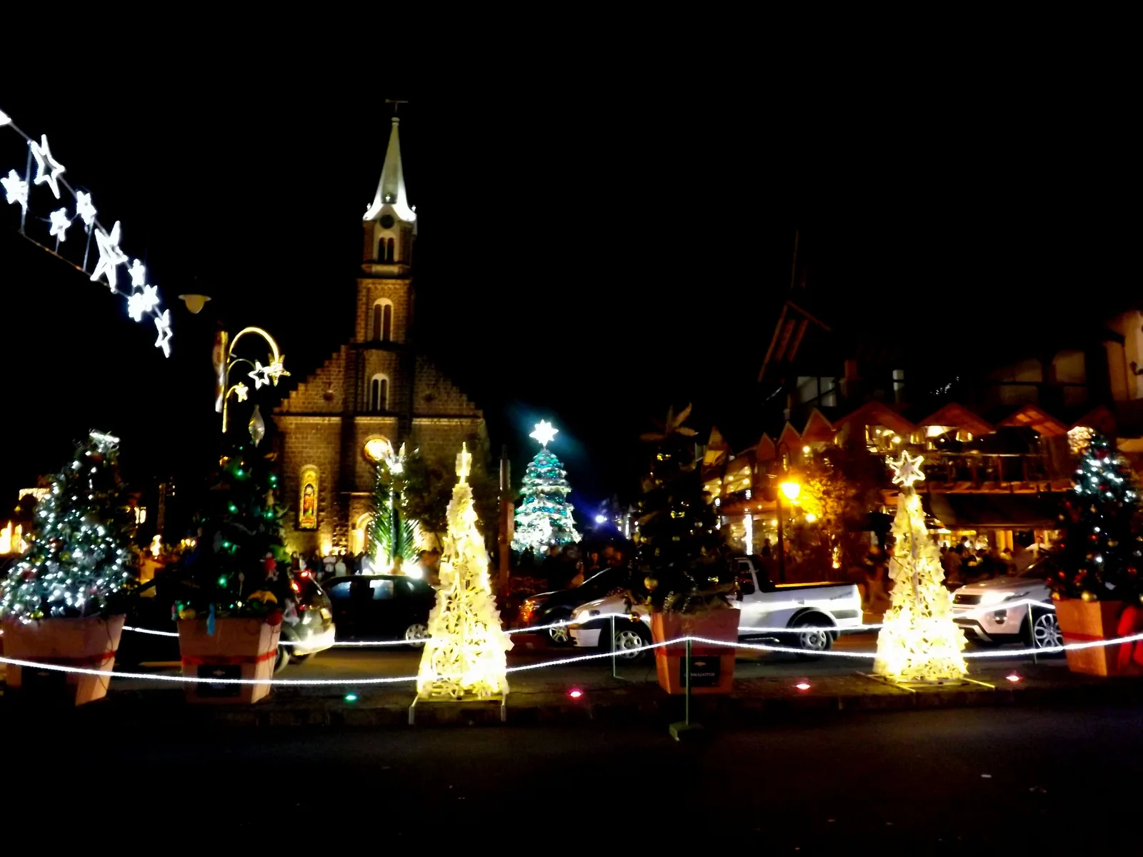 Gramado’s Igreja São Pedro at night during Natal Luz, its tower lit, with illuminated Christmas trees and strings of lights in the square in front