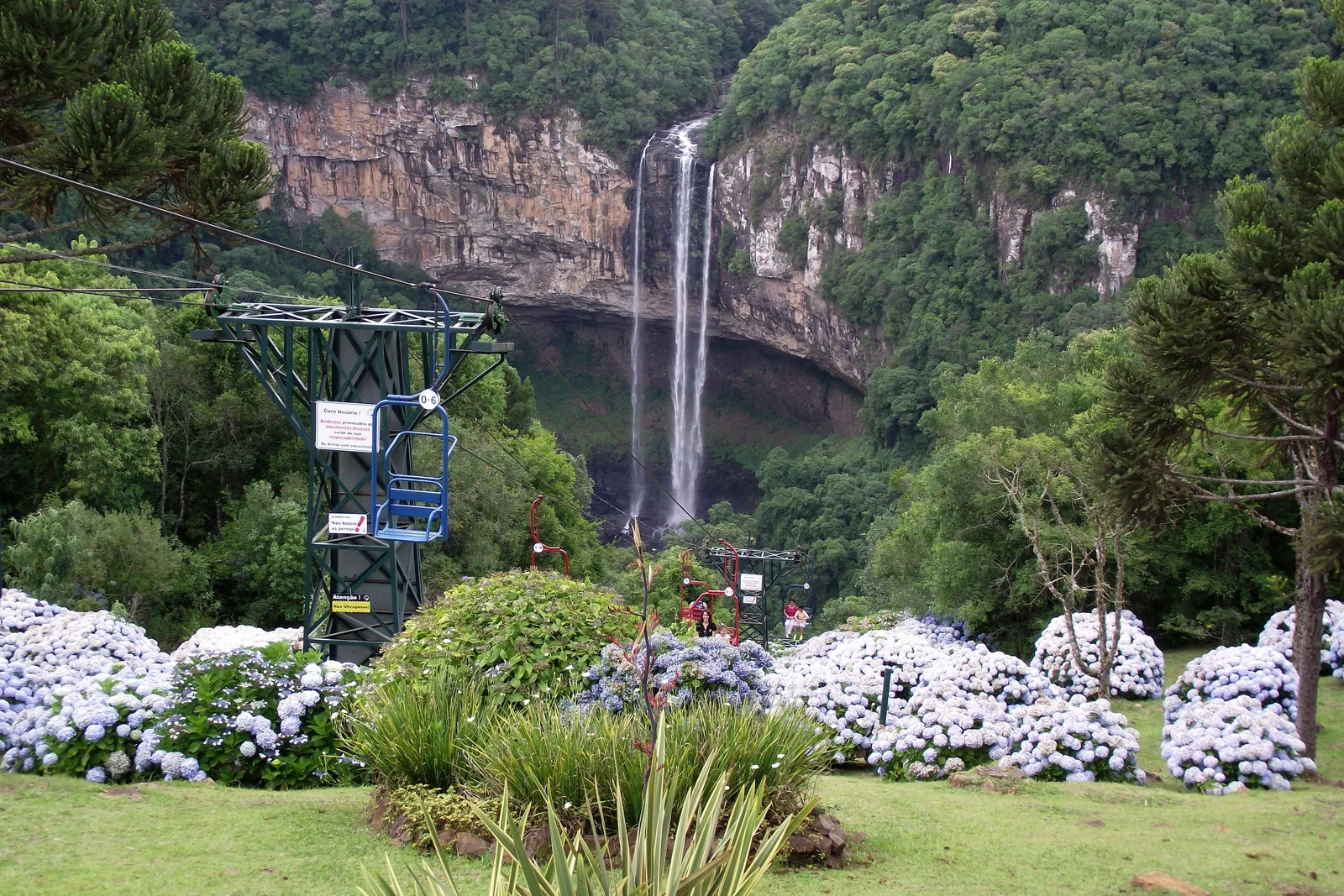 A cable-car pylon and an empty chair above beds of pale blue hydrangeas, with the Caracol waterfall falling from a cliff in the background