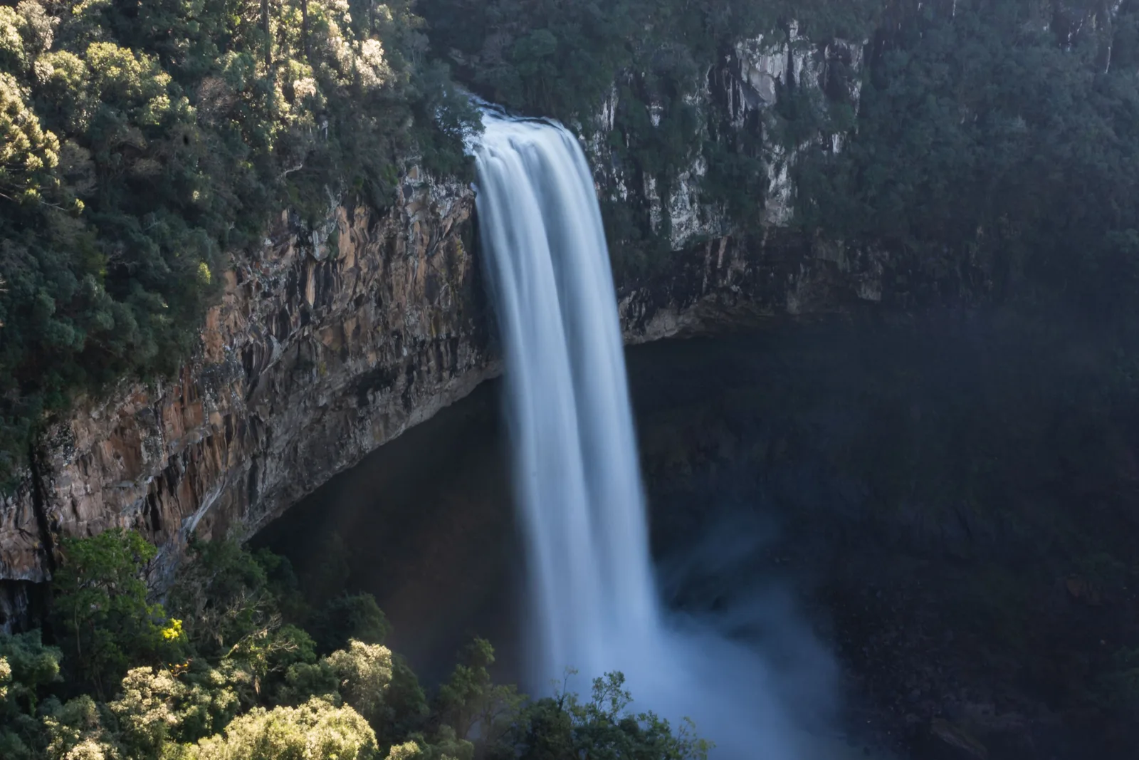 The Caracol waterfall in shadow, a bright ribbon of water dropping past a dark cliff into a misty pool, forest lit by sun along the rim