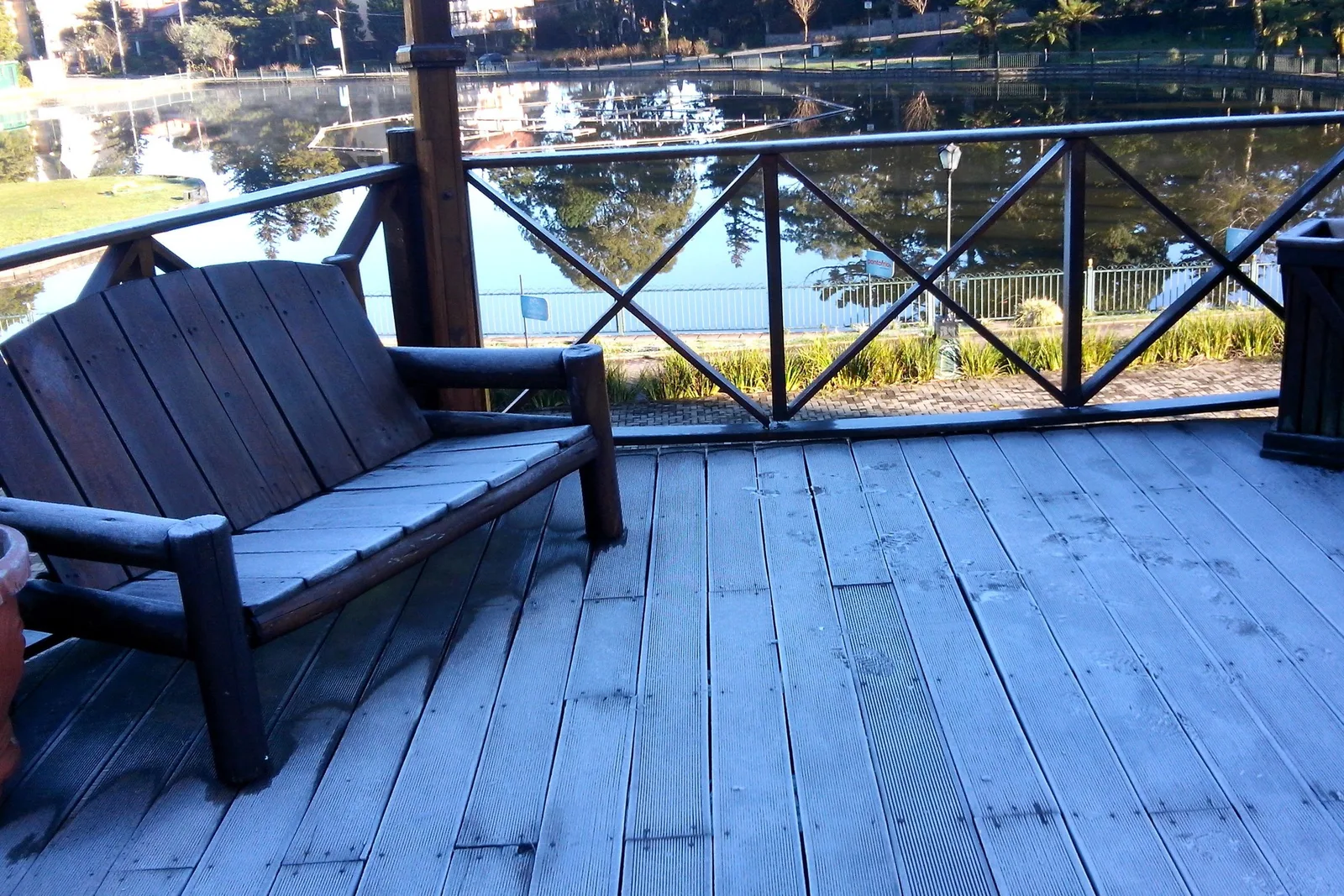 White frost on the boards and on a wooden bench of a lakeside deck, the lake and trees reflected beyond a crossed timber railing