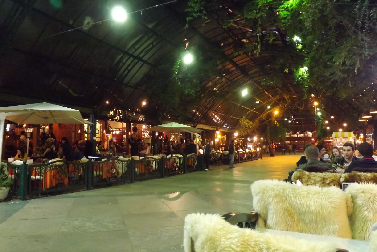 Restaurant tables under the glass roof of the Rua Coberta at night, sheepskins thrown over the chairs in the foreground and strings of lights overhead