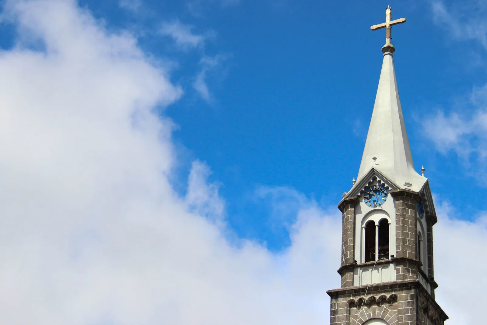 The pointed white steeple and clock of a stone church tower in Gramado against blue sky and a bank of white cloud