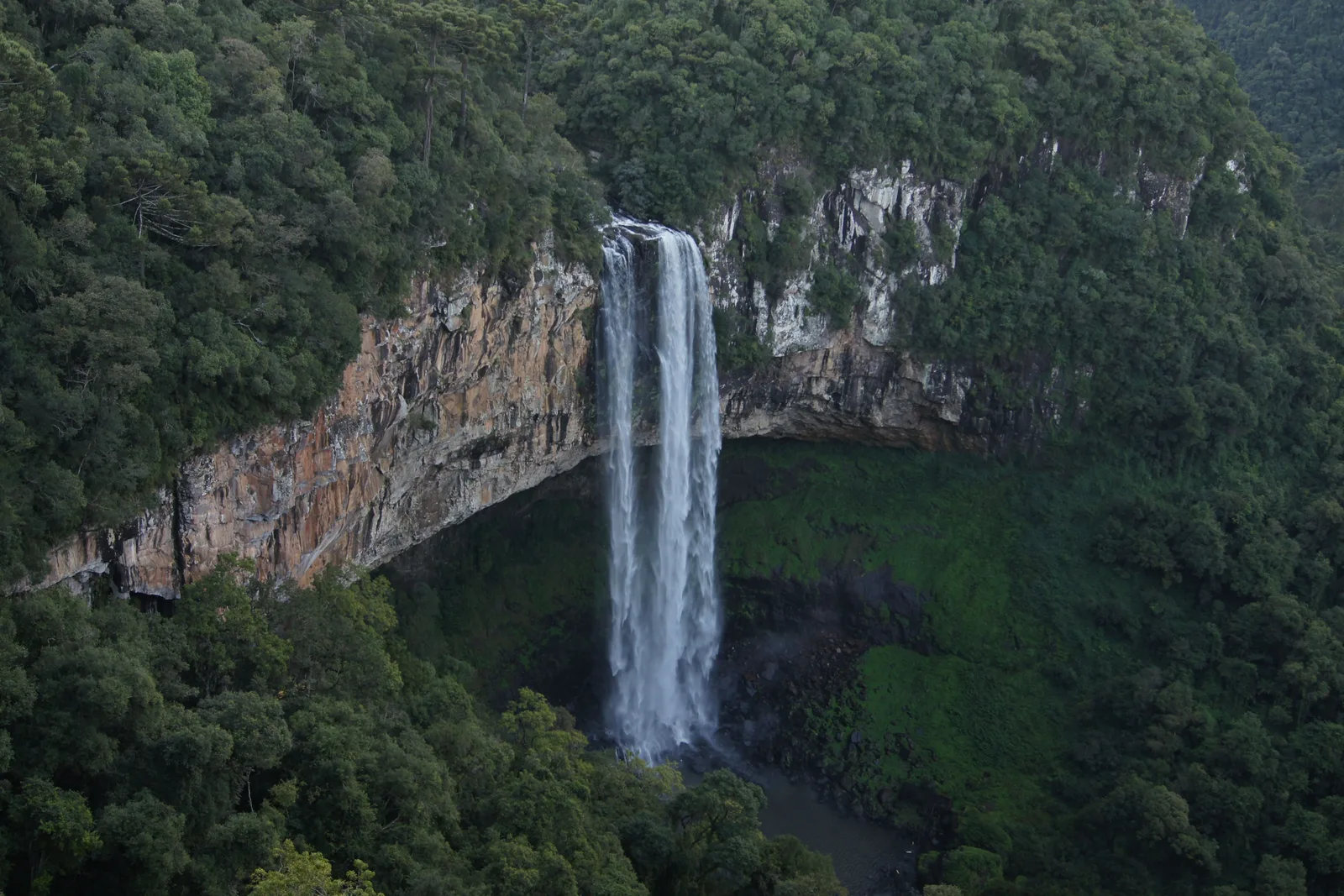 The Caracol waterfall falling from the lip of a long overhanging cliff into a forested gorge, seen across the treetops