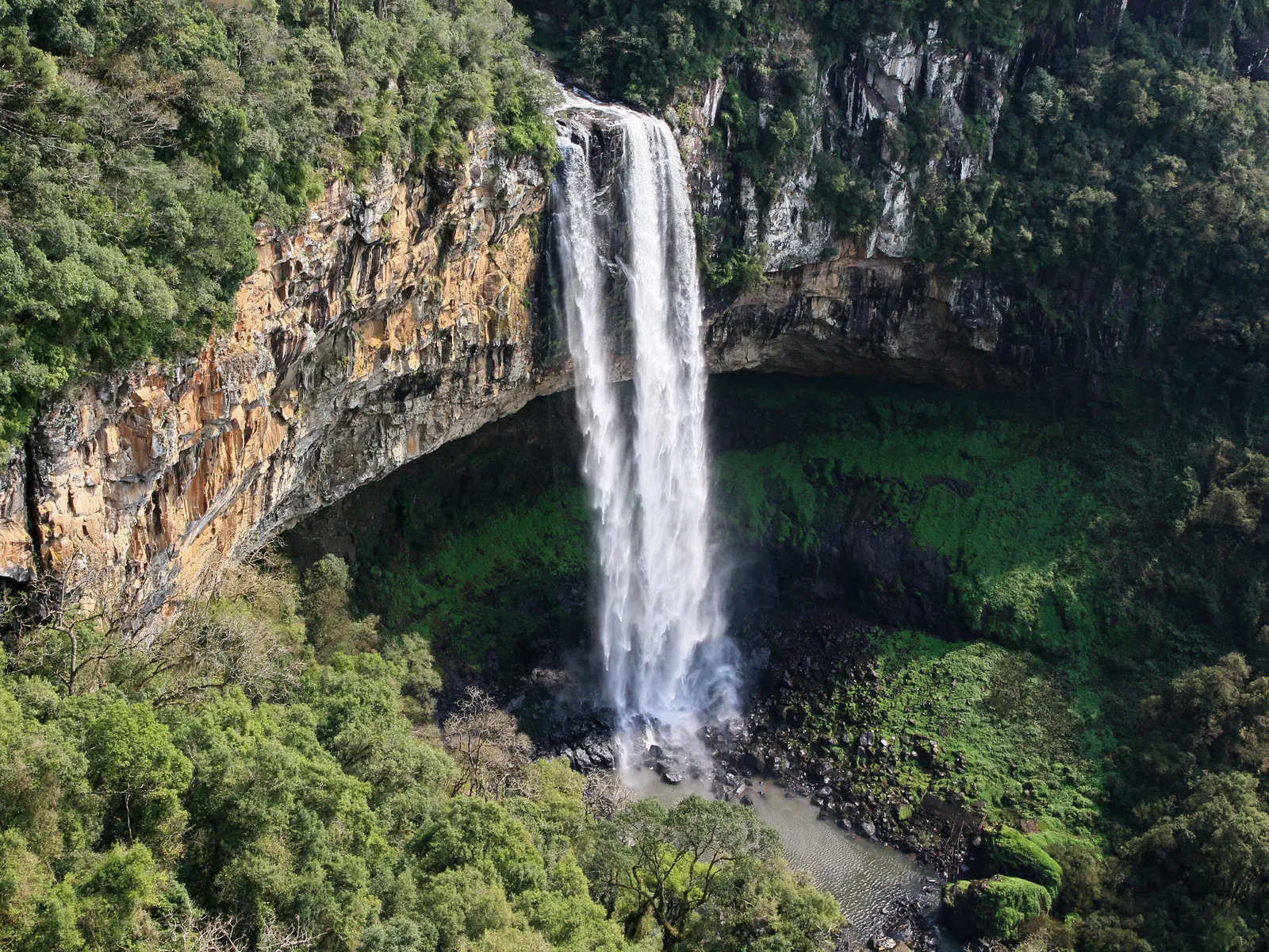 The Caracol waterfall seen from above, dropping from an overhanging orange-streaked cliff into a gorge of bright green forest, spray rising from the pool at its foot