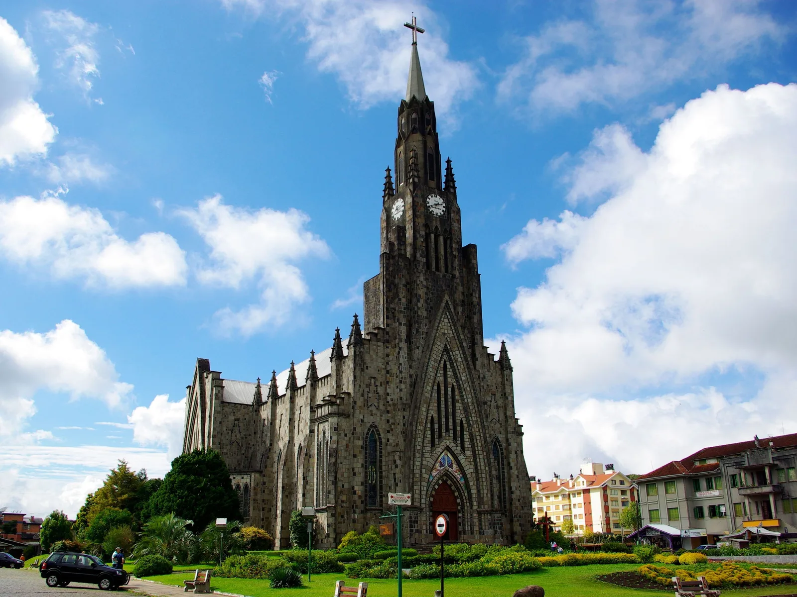 The Catedral de Pedra in Canela under a blue sky with white clouds: a dark stone Gothic church with a tall pointed clock tower, a garden and lawn in front and apartment buildings to the right