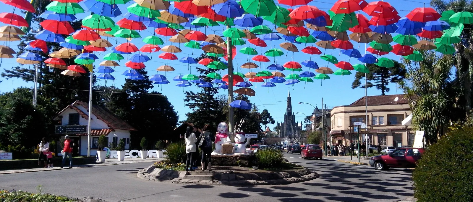 A square in central Canela under a canopy of colored umbrellas strung overhead, with the spire of the Catedral de Pedra rising at the end of the street
